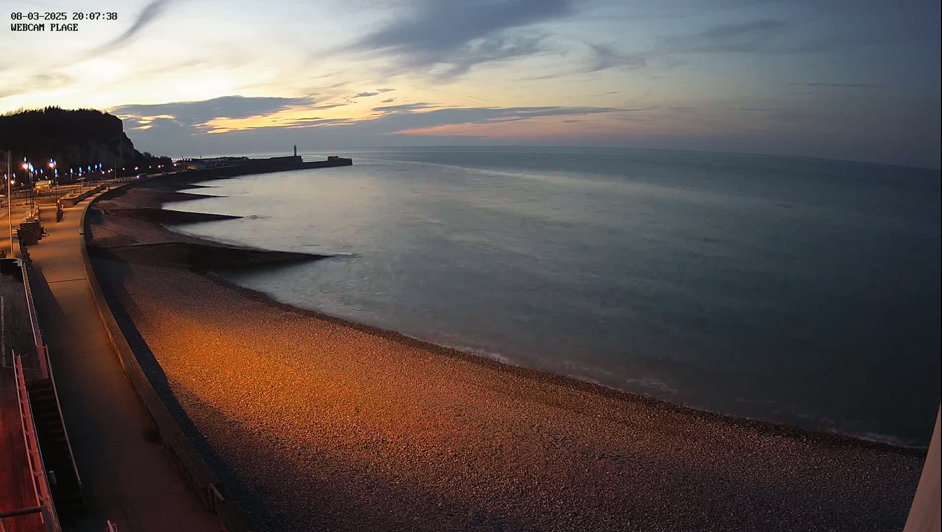 A pebble beach at twilight is illuminated by artificial lights, with calm water lapping at the shore, and a cliff visible in the distance.