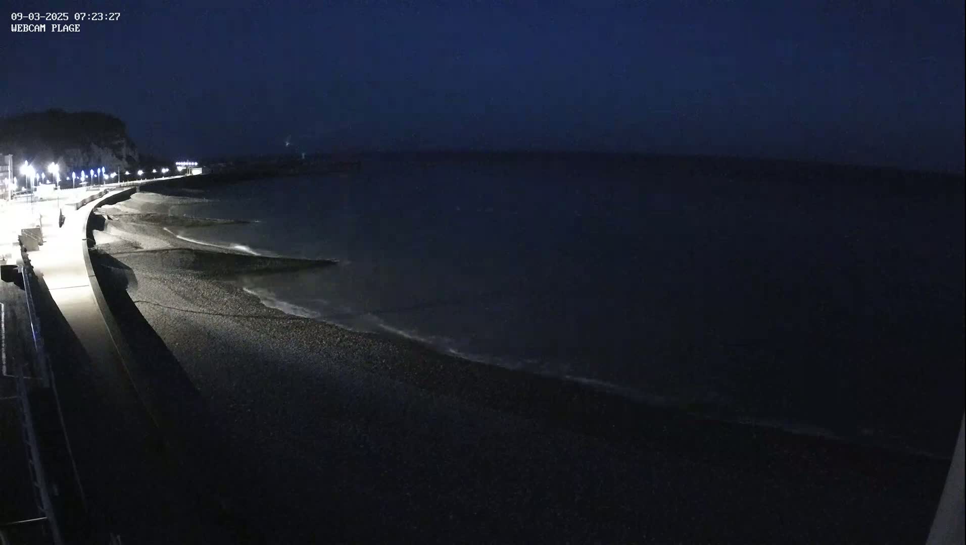 A dark, nighttime view of a pebble beach and a paved walkway beside a body of water under a clear, dark sky.