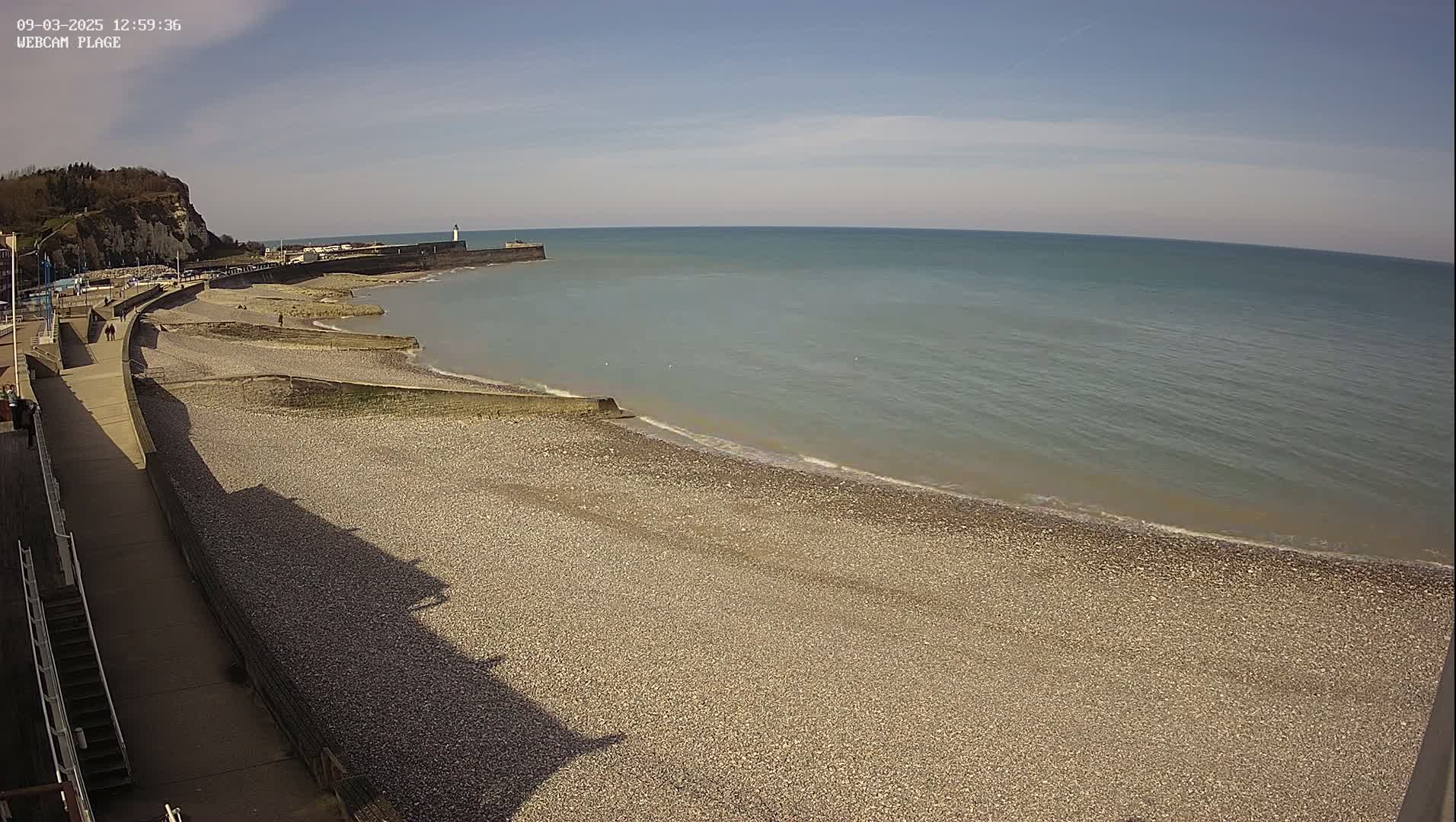 A pebble beach curves around a calm, light-blue sea under a clear, sunny sky, next to a paved walkway and a cliff in the background.