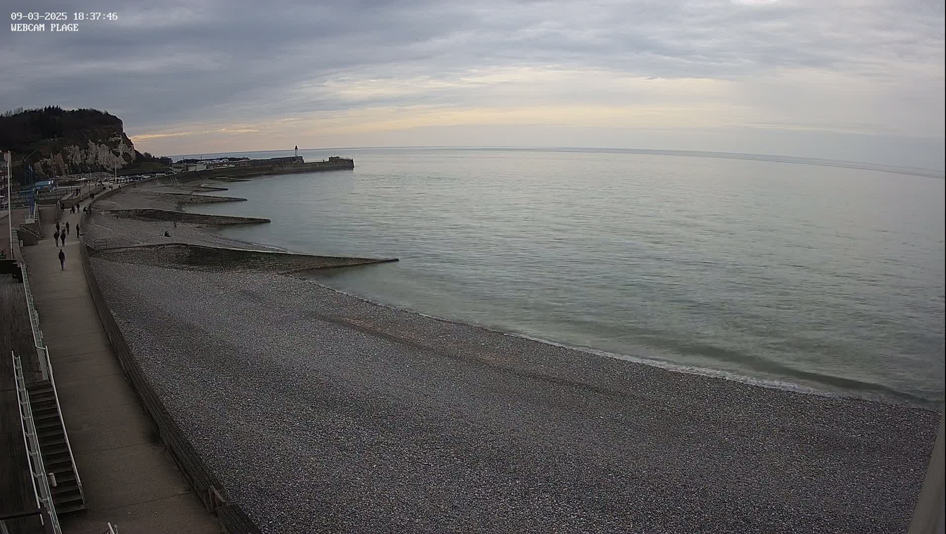 A pebble beach stretches alongside a paved walkway under an overcast sky, with a calm sea extending to the horizon and a distant cliff face visible in the background.