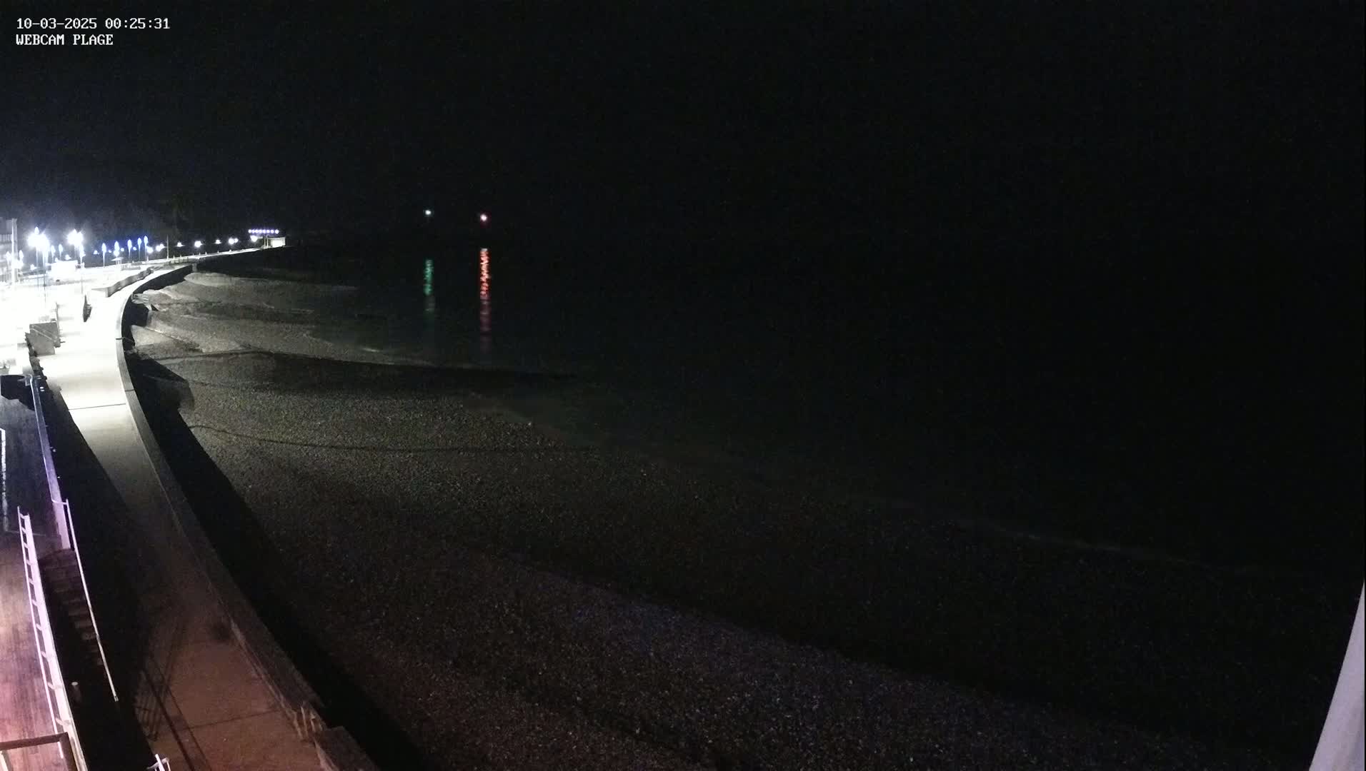A dark, nighttime view of a pebble beach and a paved walkway, illuminated by streetlights, with calm water visible in the background.
