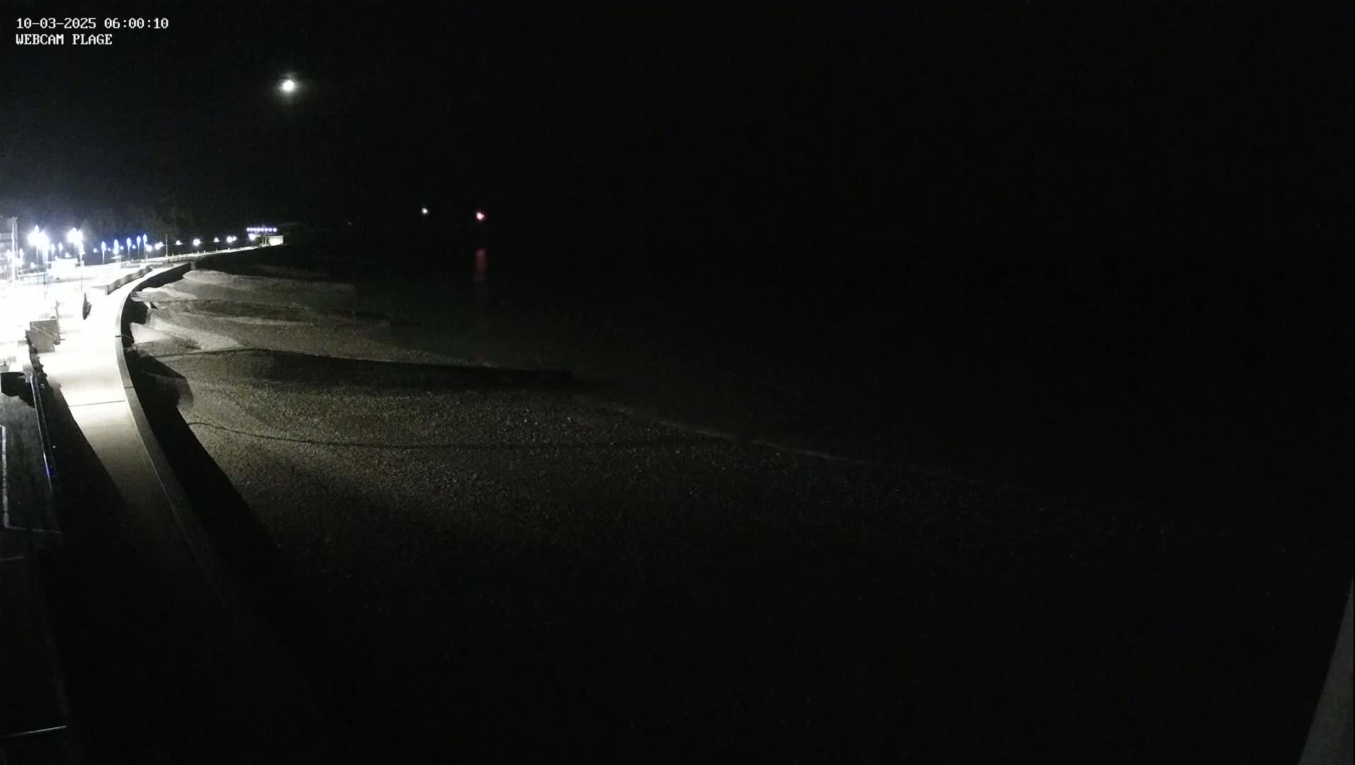 A dark, moonlit night scene shows a paved walkway curving alongside a dark, pebble beach where gentle waves lap the shore.