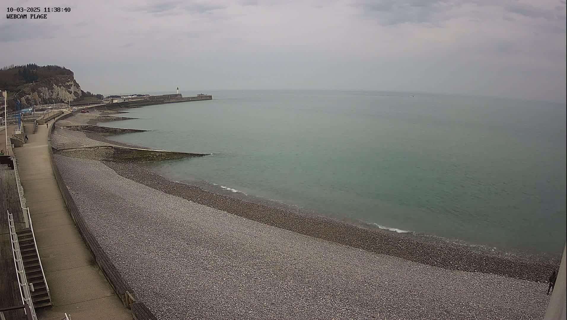 A pebbled beach and a paved walkway curve alongside a calm, greenish-grey sea under an overcast sky.