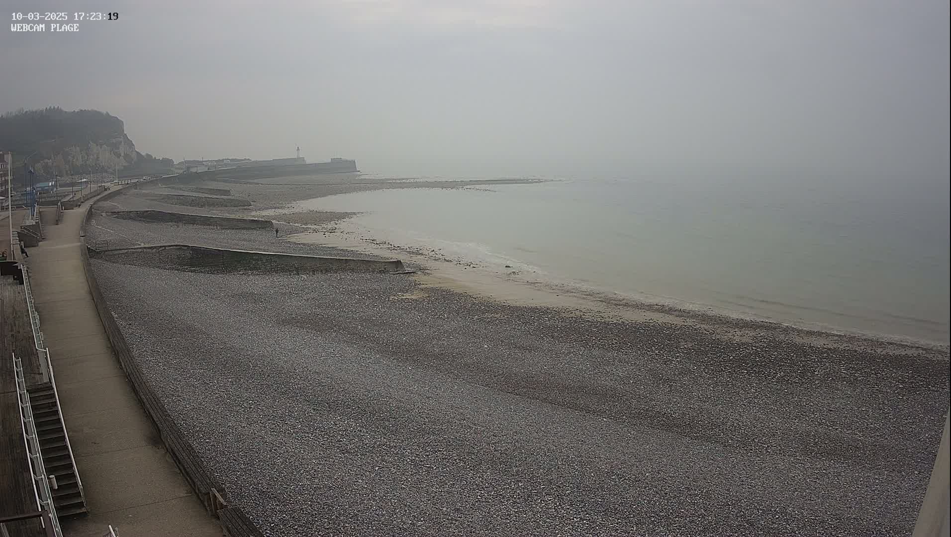 A pebble beach stretches along a calm, foggy sea, backed by a paved walkway and a cliff.