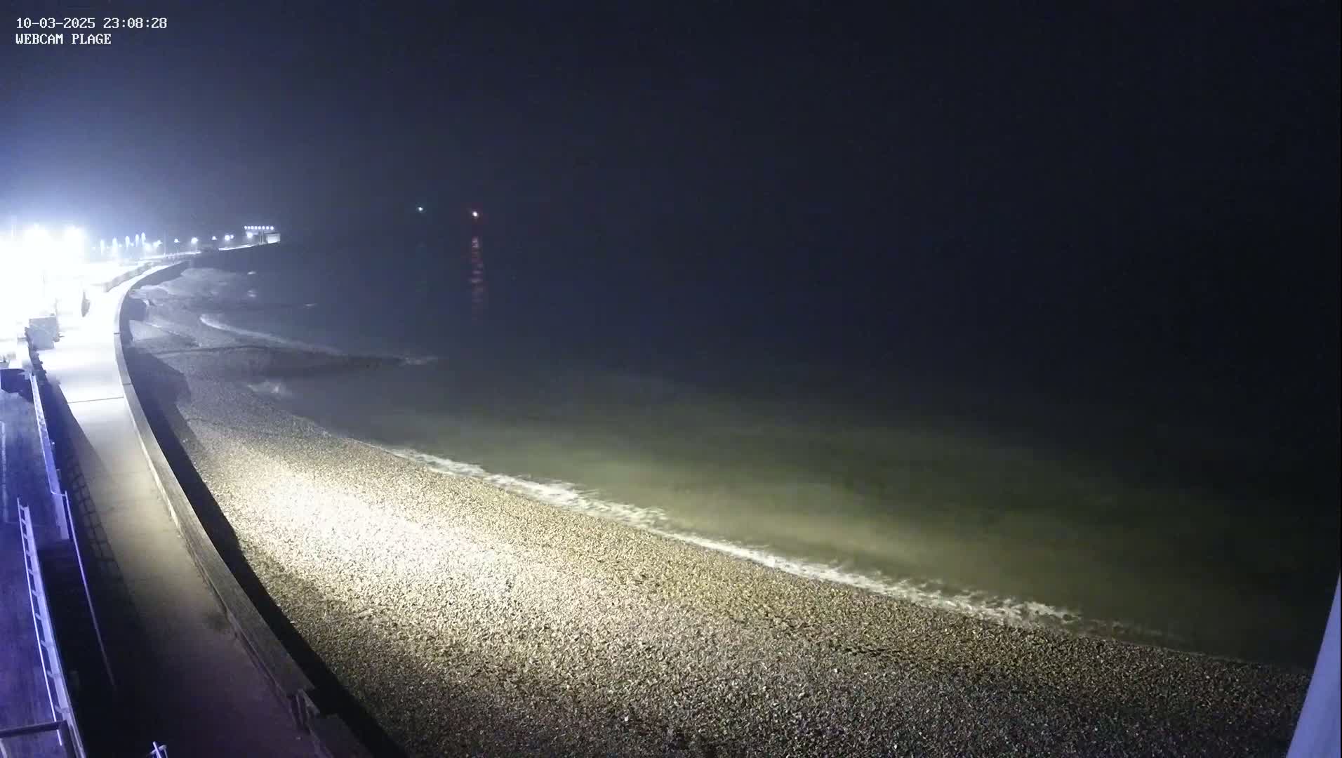 A nighttime, hazy view of a pebble beach and a paved walkway next to the ocean, with gentle waves lapping the shore.