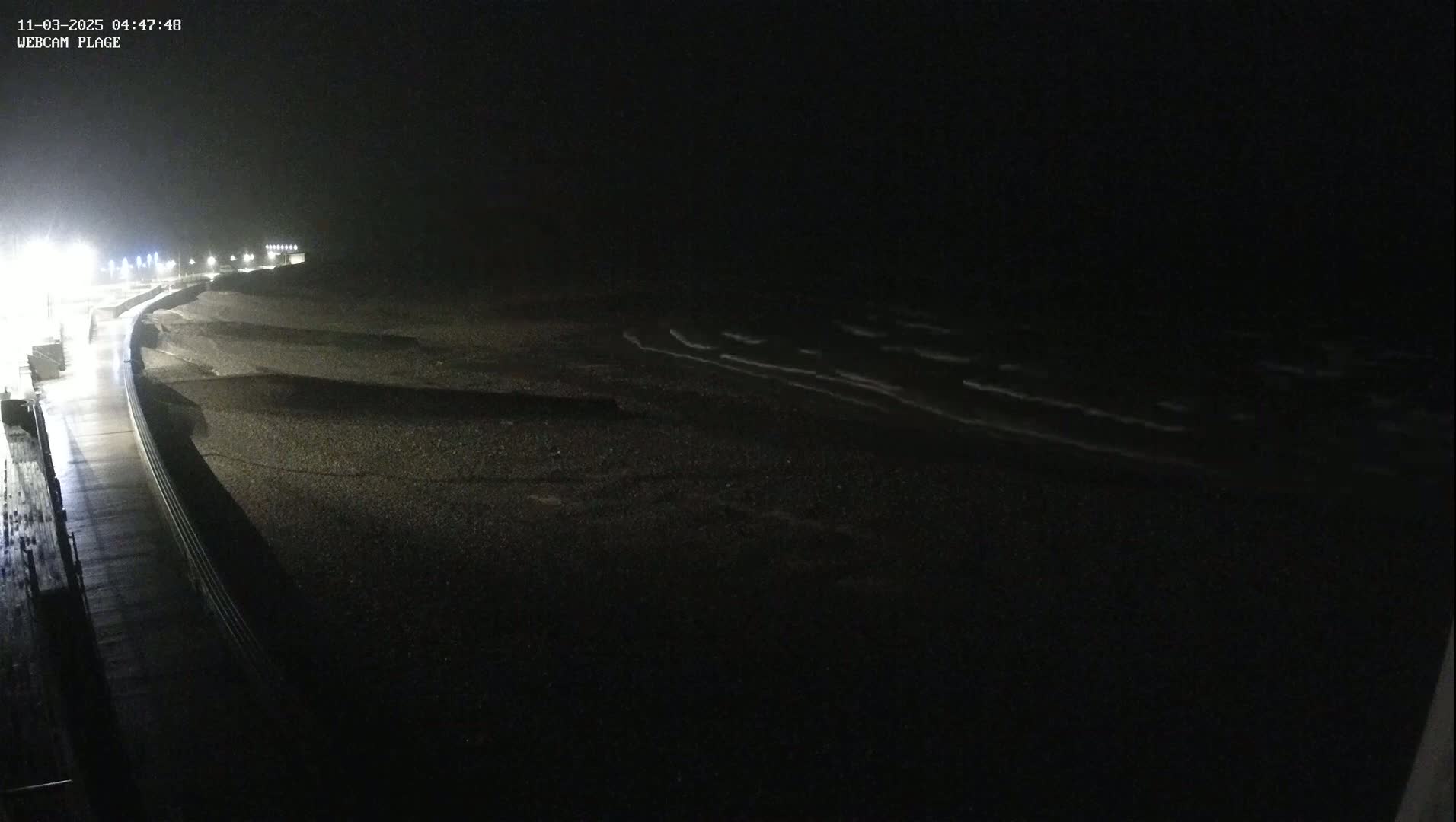 A dark, nighttime view of a beach with gently lapping waves under a mostly clear sky, showing a wet roadway and a seawall next to the beach.