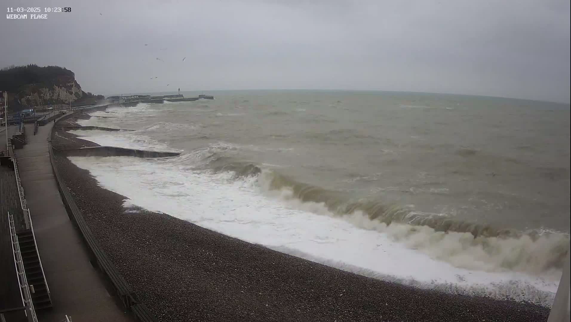 Large waves crash against a pebble beach next to a paved walkway and cliff on an overcast day.