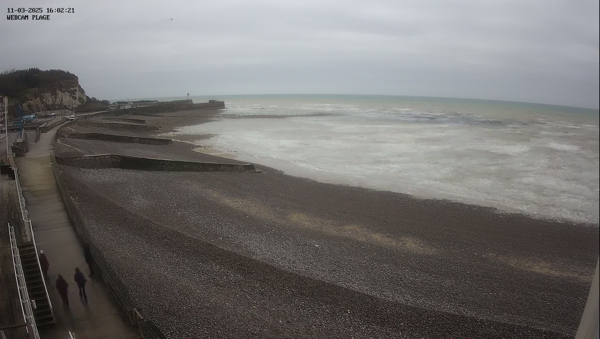 A pebble beach stretches out to a calm, grey sea under an overcast sky, with a paved walkway and a few people walking along the shore.