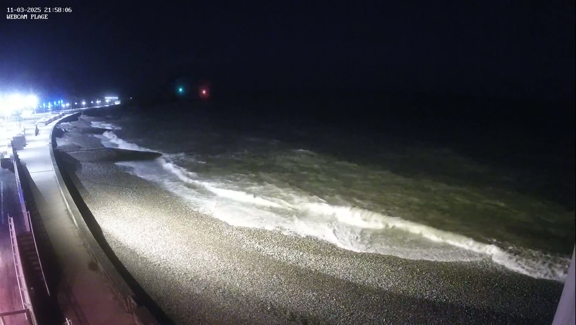 A dark, nighttime beach scene shows waves lapping onto a pebbled shore next to a paved walkway with lights.