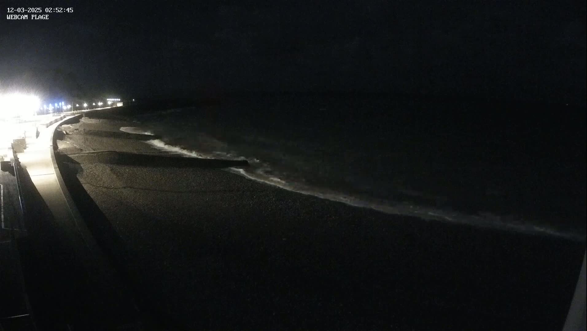 A dark, nighttime beach scene shows gently lapping waves on a pebble shore next to a dimly lit walkway.