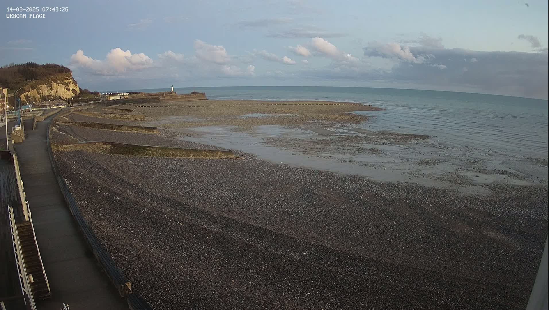 A mostly cloudy day reveals a pebble beach with a walkway and a calm sea stretching to a cliffside in the background.