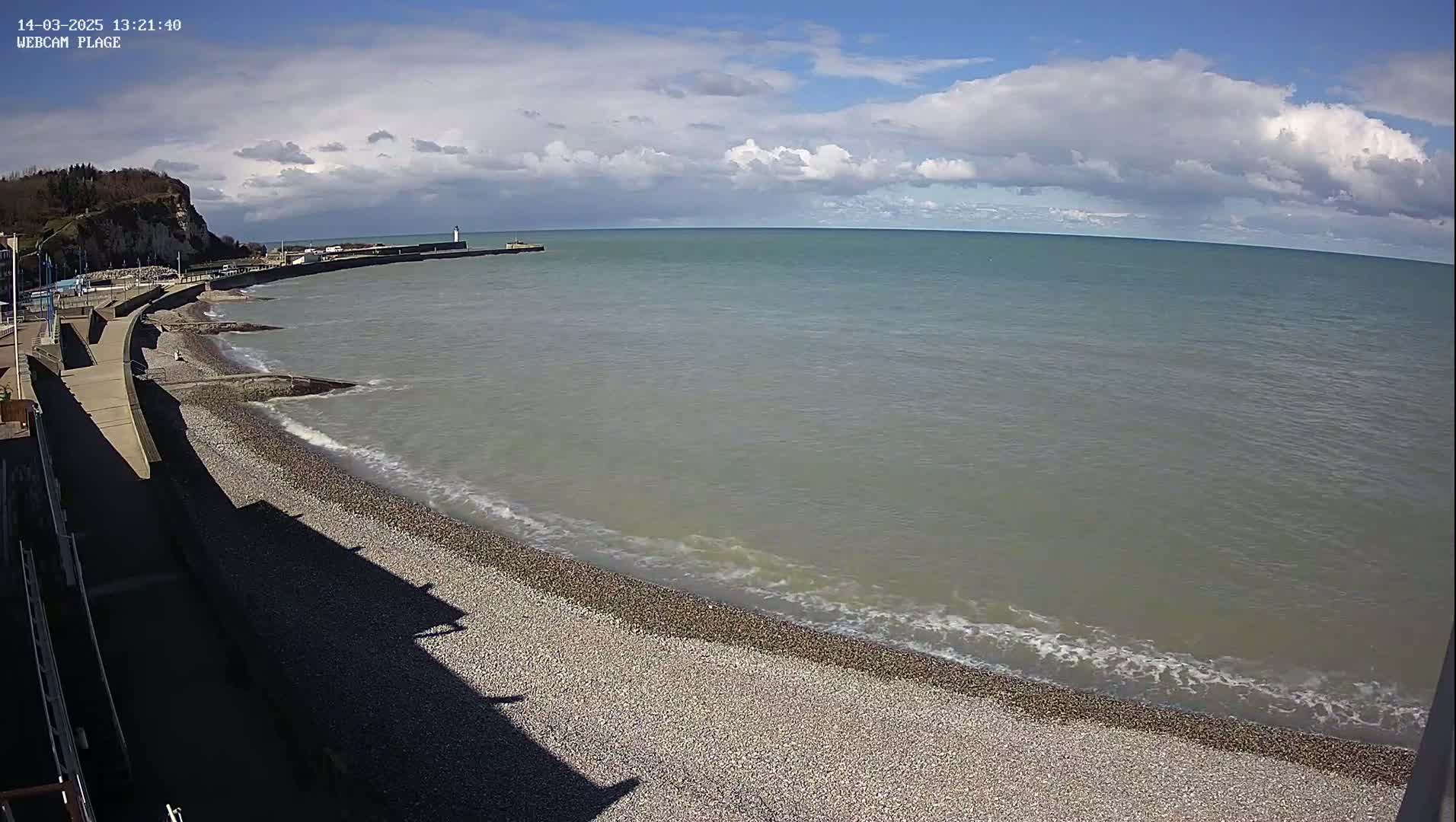 A pebble beach curves along a calm sea under a partly cloudy sky, with a cliff and buildings visible in the distance.