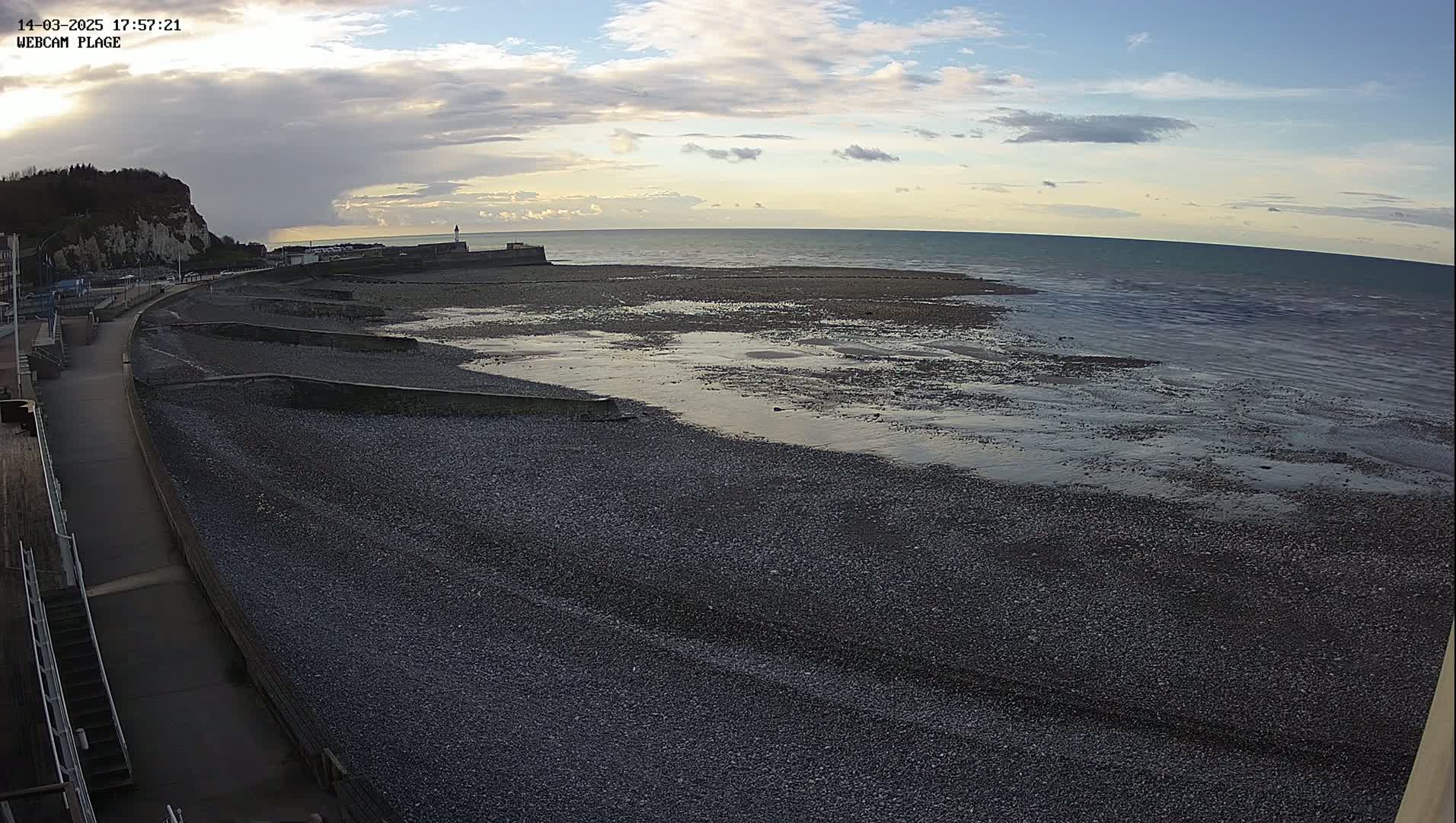 A pebble beach stretches out to a calm sea under a partly cloudy sky at sunset.