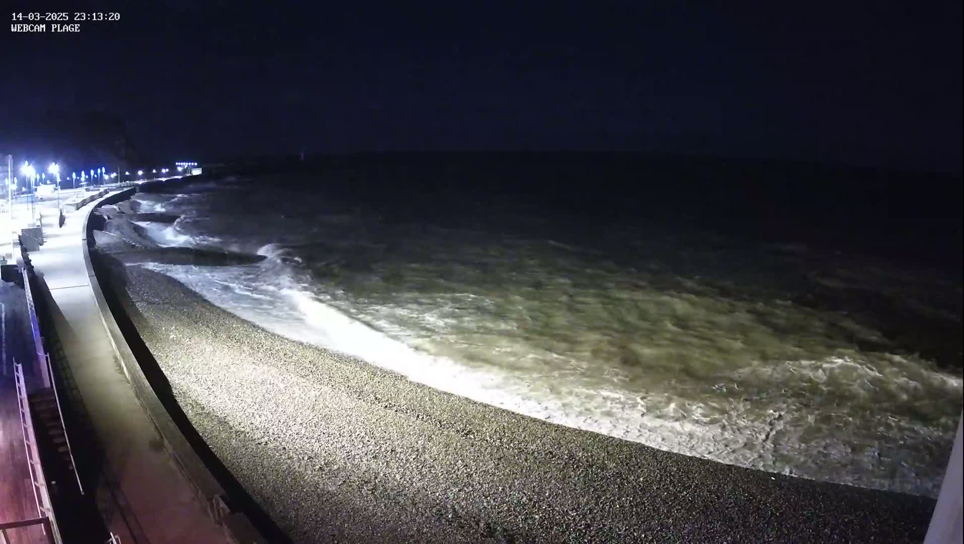 A nighttime coastal scene shows moderately rough waves crashing on a pebble beach next to a lit walkway.