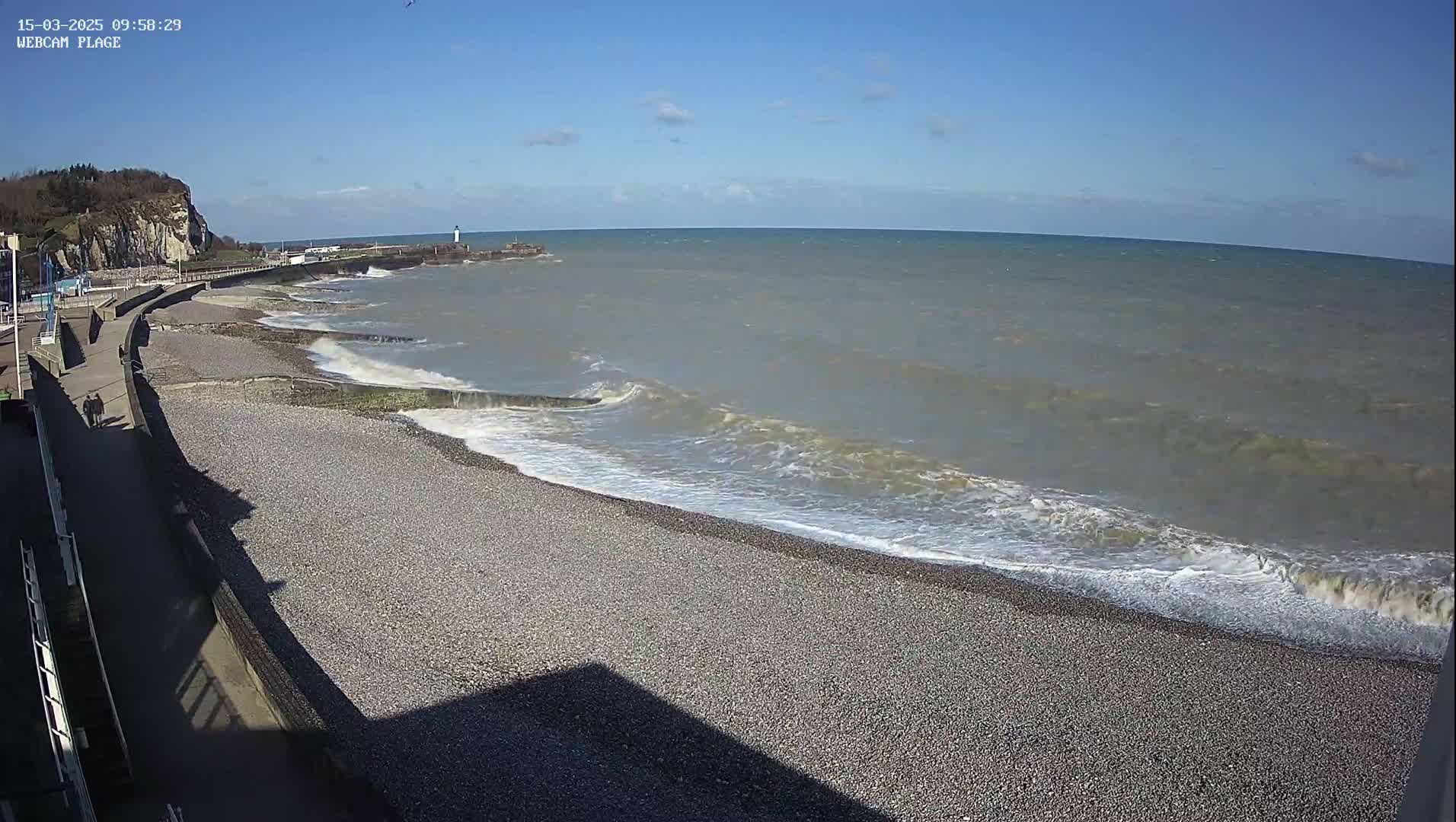 A rocky beach with small waves rolling onto the shore on a sunny day, with a walkway and cliffs visible in the background.