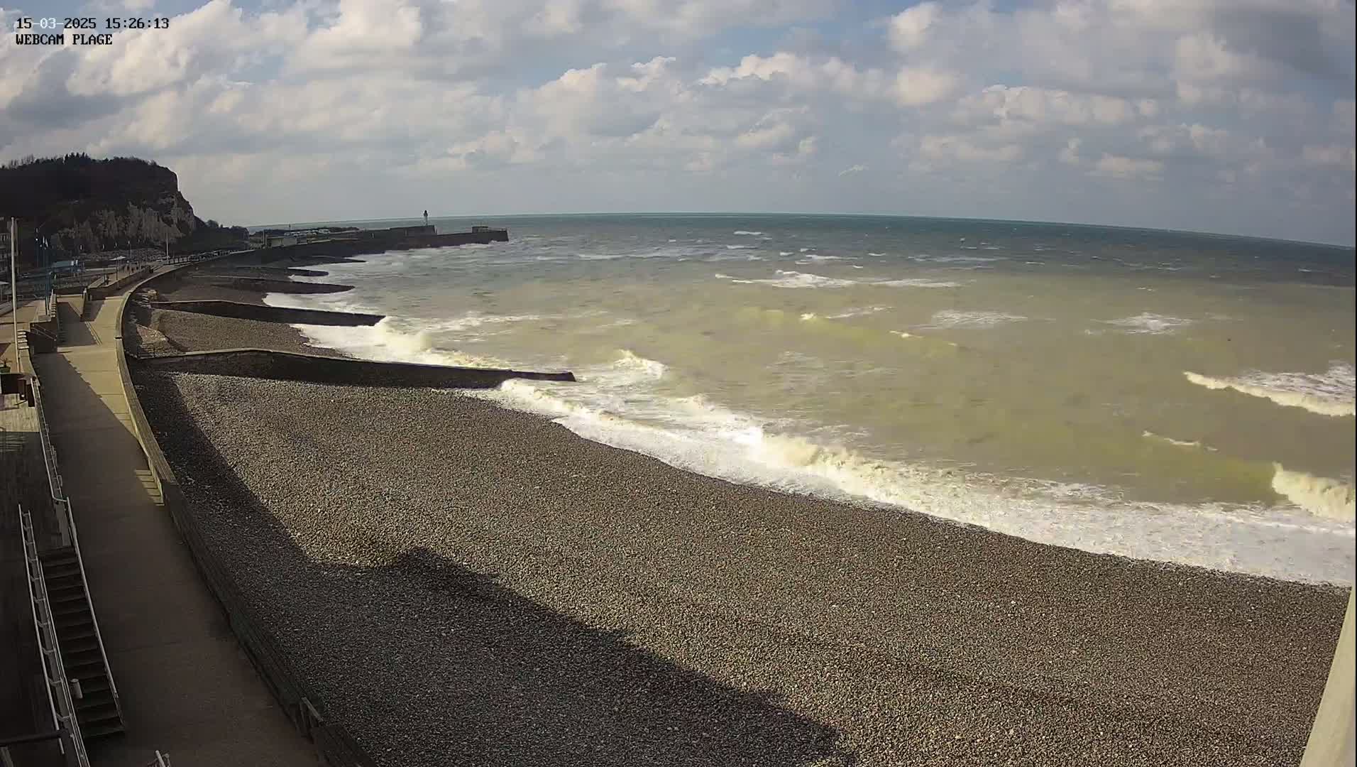 A pebble beach with a walkway and sea wall is shown with moderately sized waves breaking on the shore under a partly cloudy sky.
