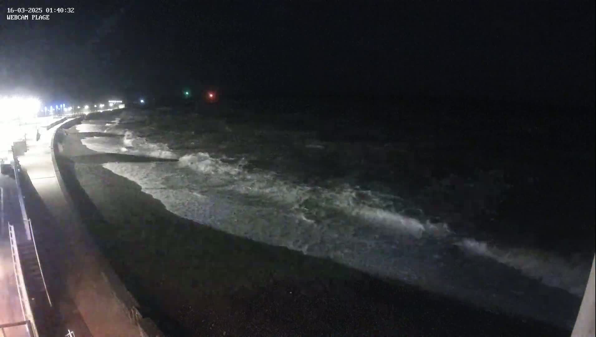 At night, moderately sized waves crash on a dark, pebbly beach next to a paved walkway and dimly lit shoreline.
