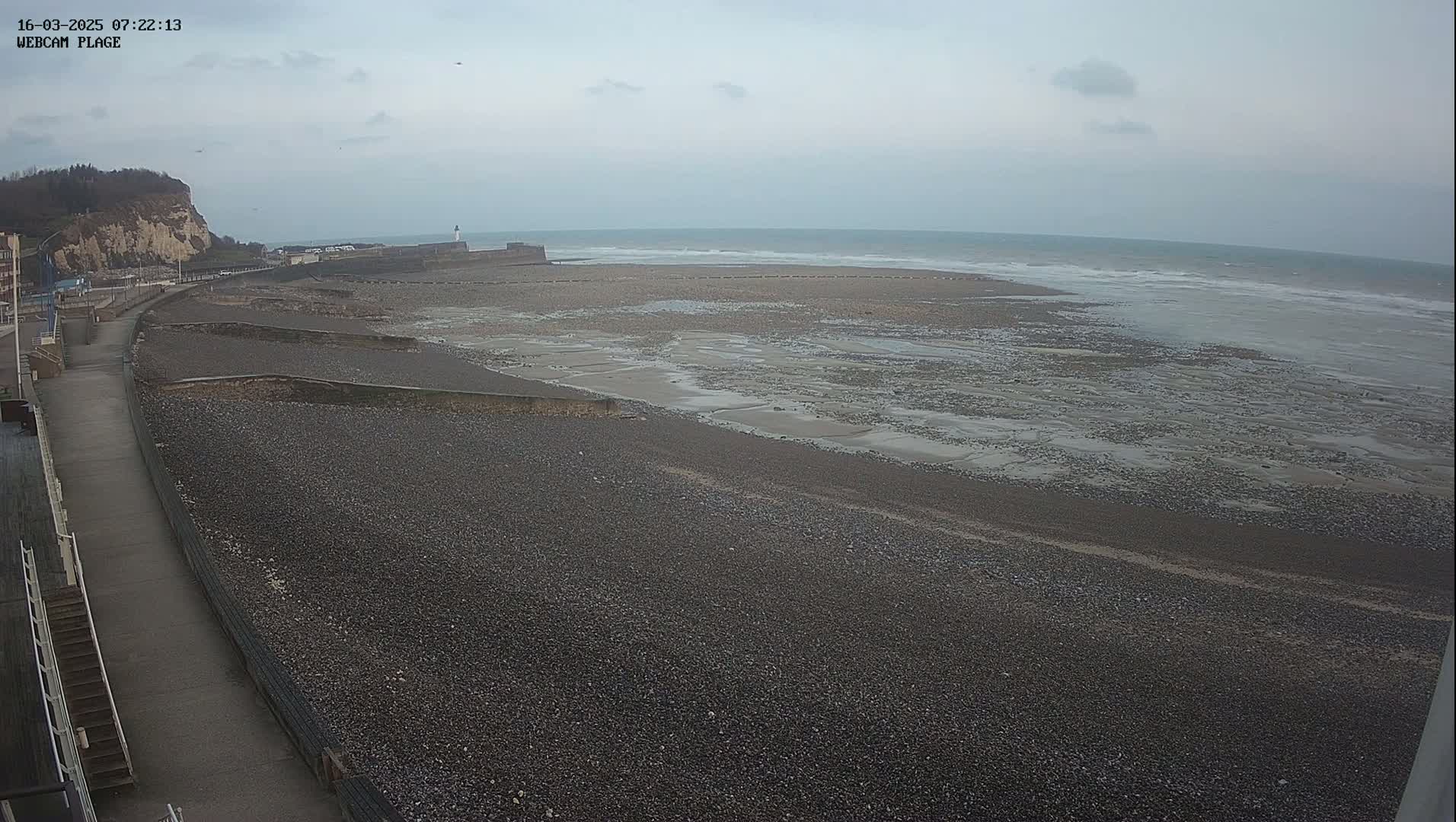 A pebble beach stretches out to a calm, gray sea under an overcast sky, with a paved walkway and a cliff visible in the background.