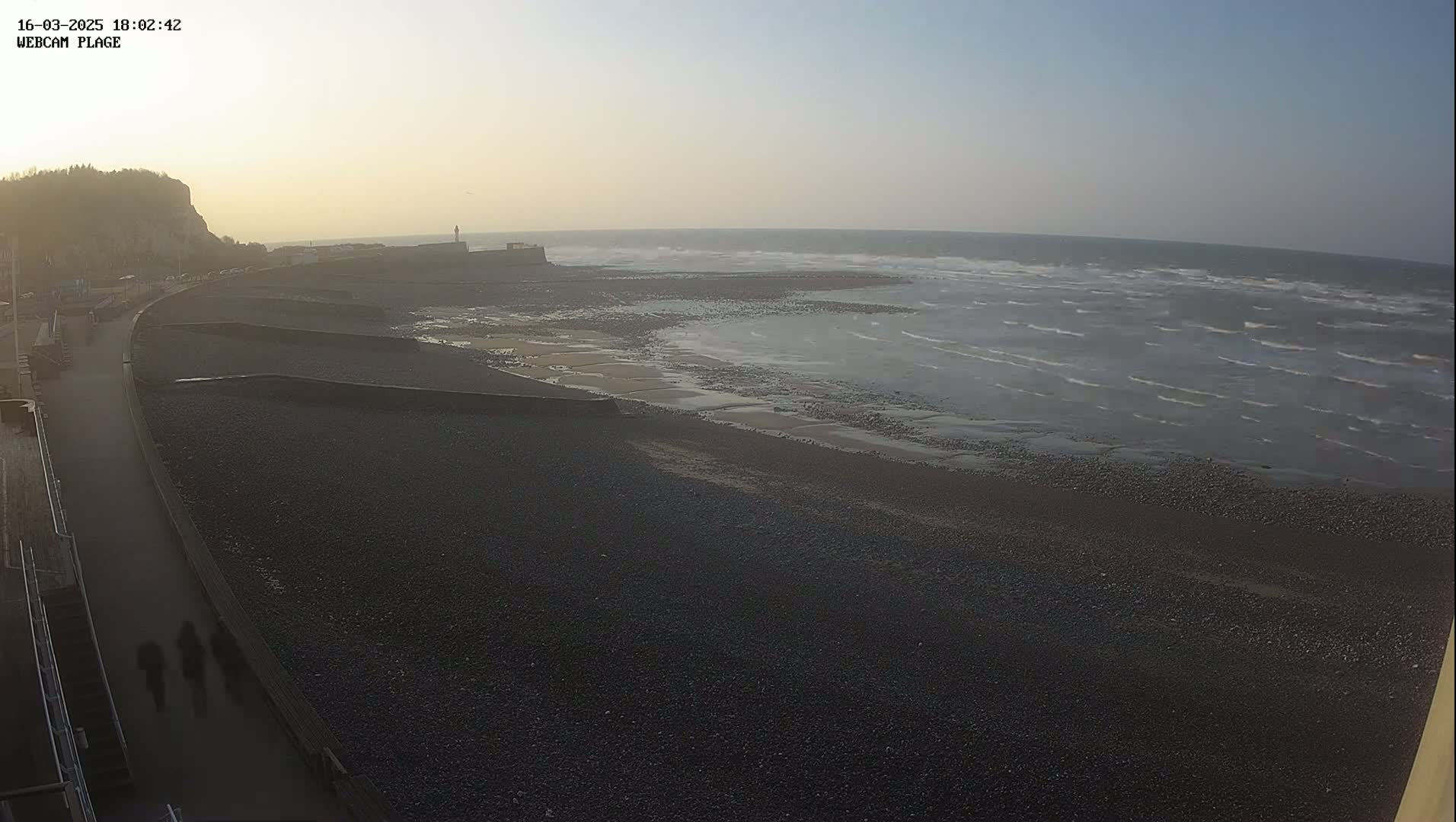 A dark, pebbly beach stretches along a paved walkway, under a hazy, light sky, with small waves rolling into the shore.