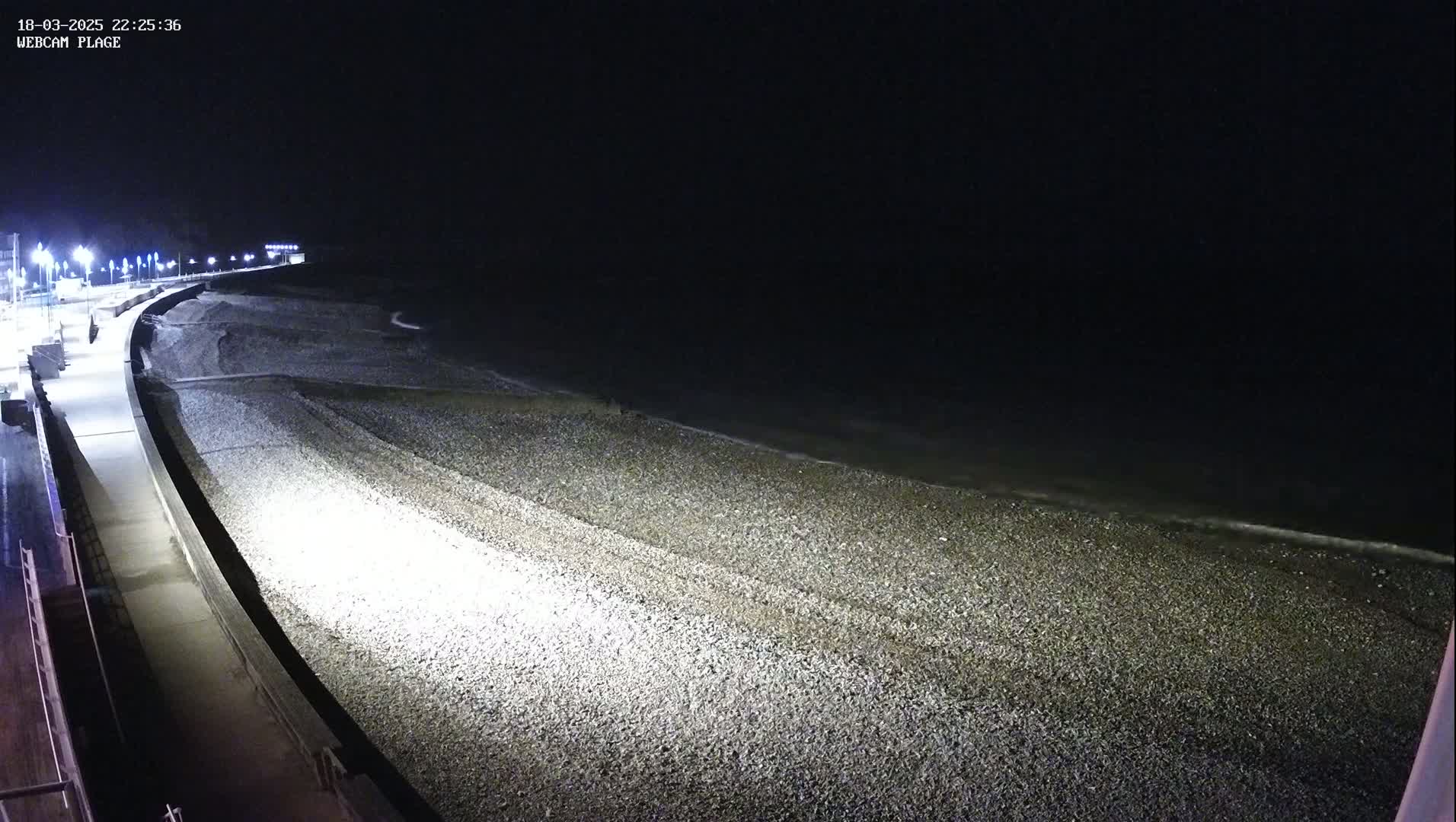 A dark, nighttime view of a pebble beach and adjacent paved walkway, illuminated by artificial lights, with a calm sea in the background.