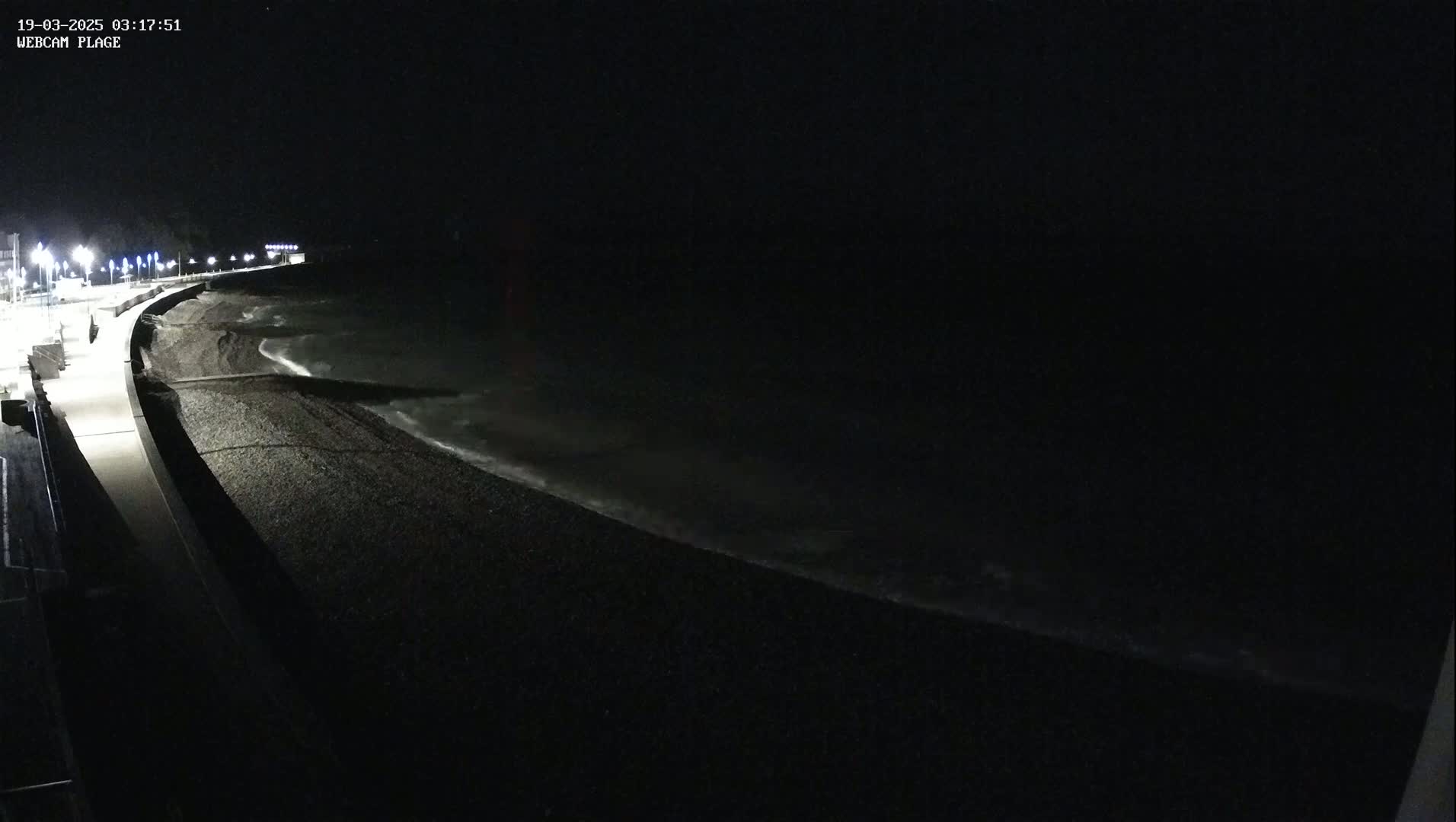 A dark, nighttime view of a pebble beach with gentle waves lapping the shore, illuminated by streetlights along a nearby paved walkway.