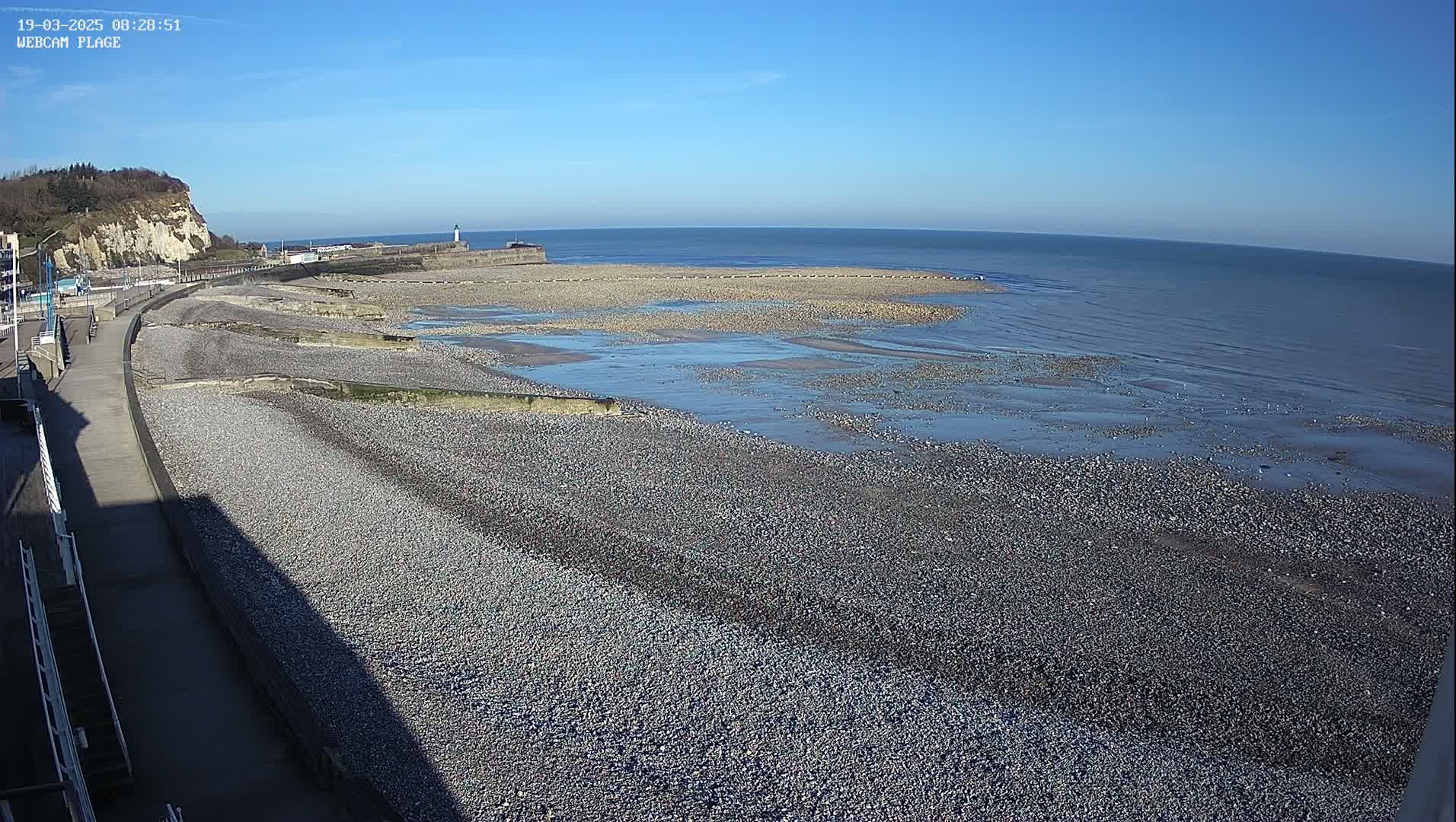 A pebble beach stretches along a calm, blue sea under a clear blue sky, with a paved walkway and a cliff visible in the background.