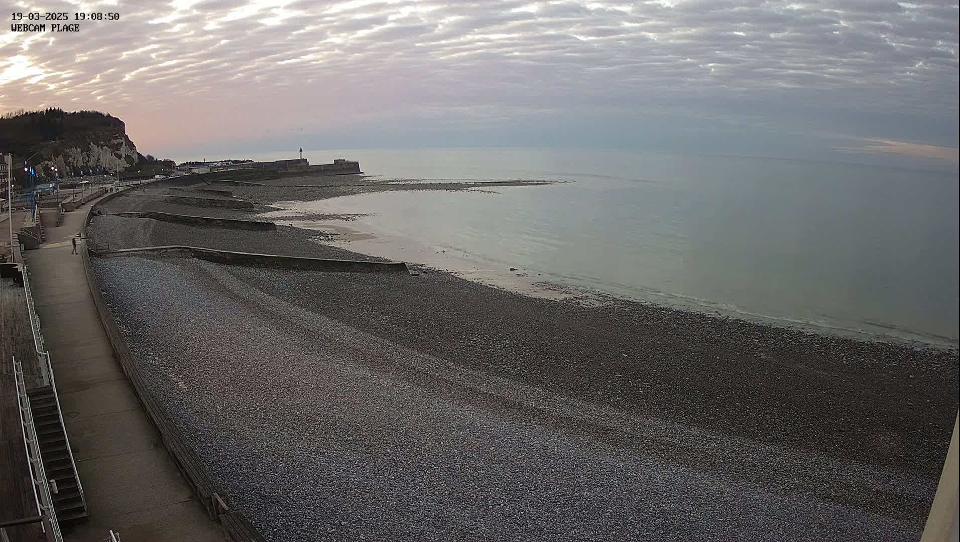 A rocky beach with a seawall and a paved walkway extends from a cliffside town under a cloudy, overcast sky.