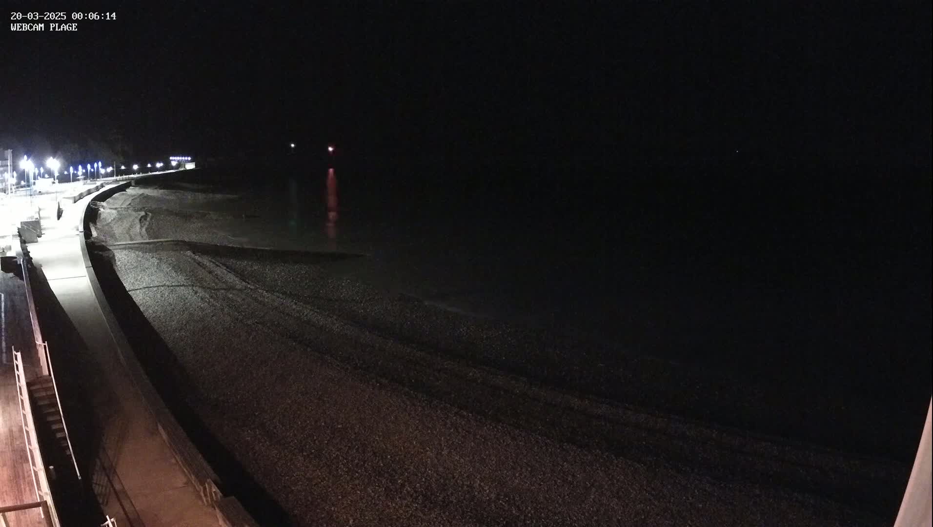 A dark, pebble beach is shown at night, illuminated by streetlights along a paved walkway.