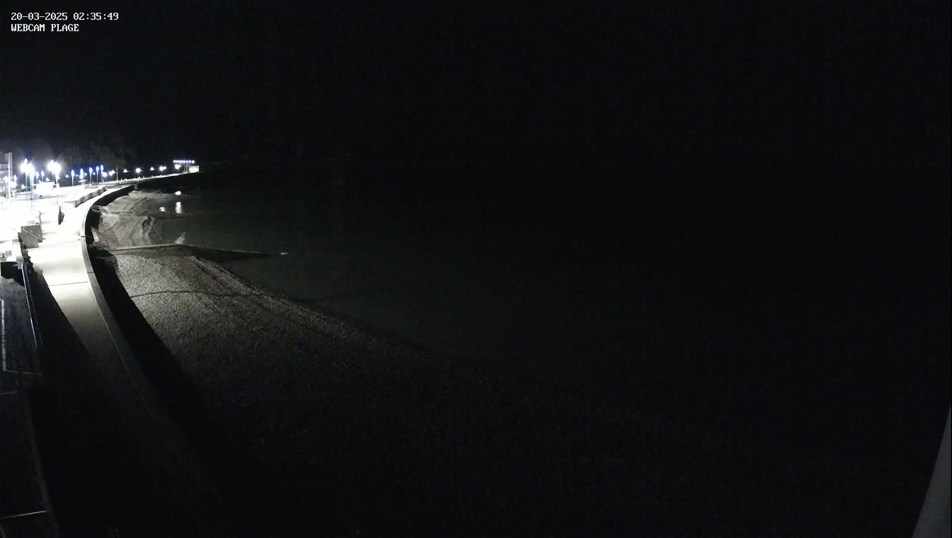 A dark, nighttime view shows a paved walkway curving along a pebble beach and a dark body of water under a clear, black sky.