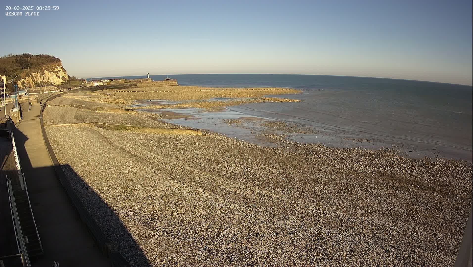 A pebble beach stretches along a calm sea under a clear blue sky, with a paved walkway and a cliff visible in the background.