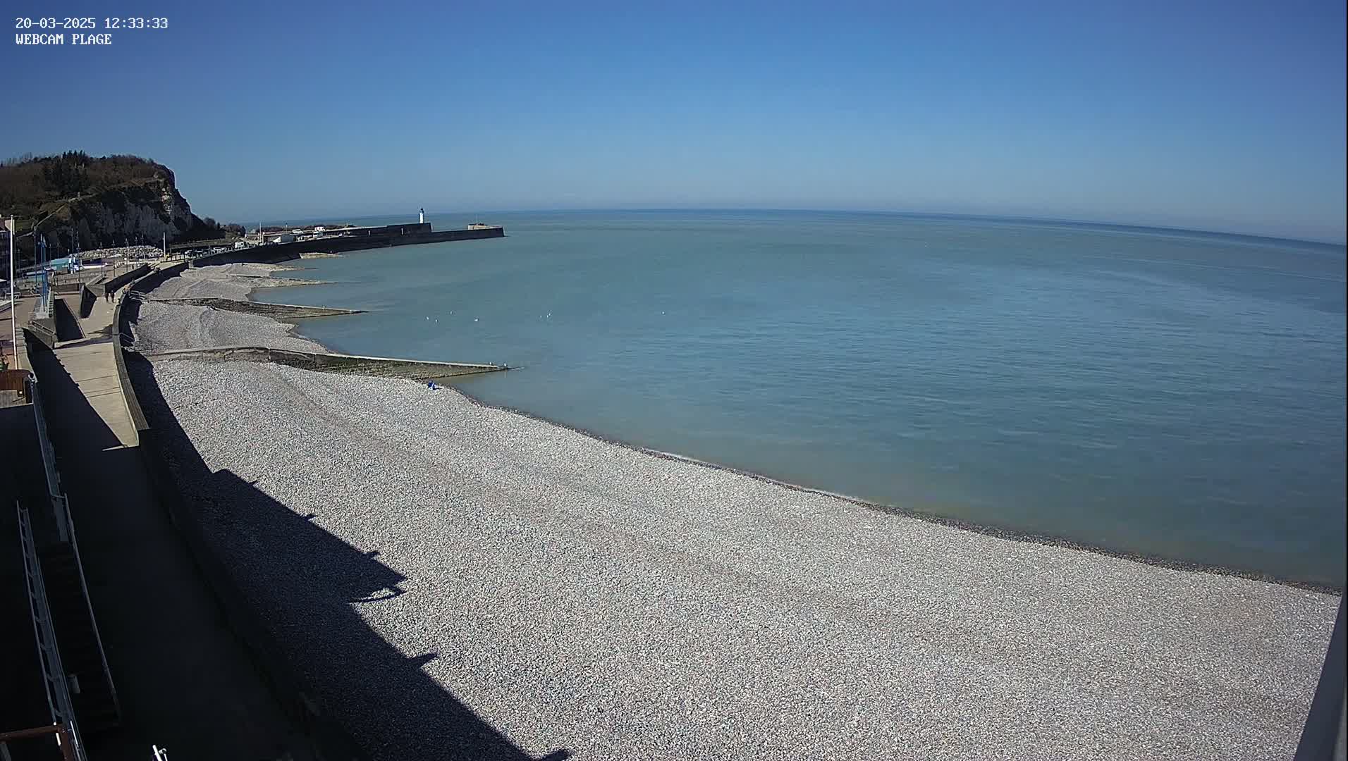 A pebble beach curves along a calm, blue ocean under a clear, sunny sky, with a cliff and seawall visible in the distance.