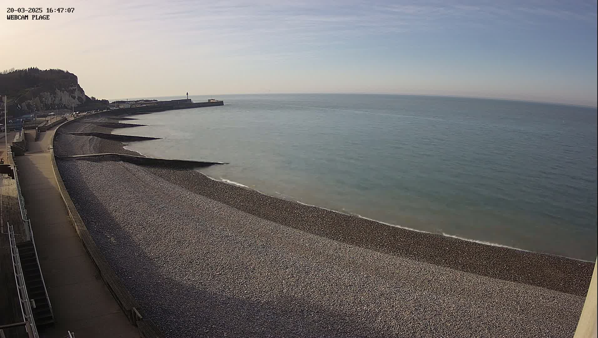 A pebble beach stretches along a calm sea under a clear, bright sky, next to a paved walkway and a cliff in the distance.