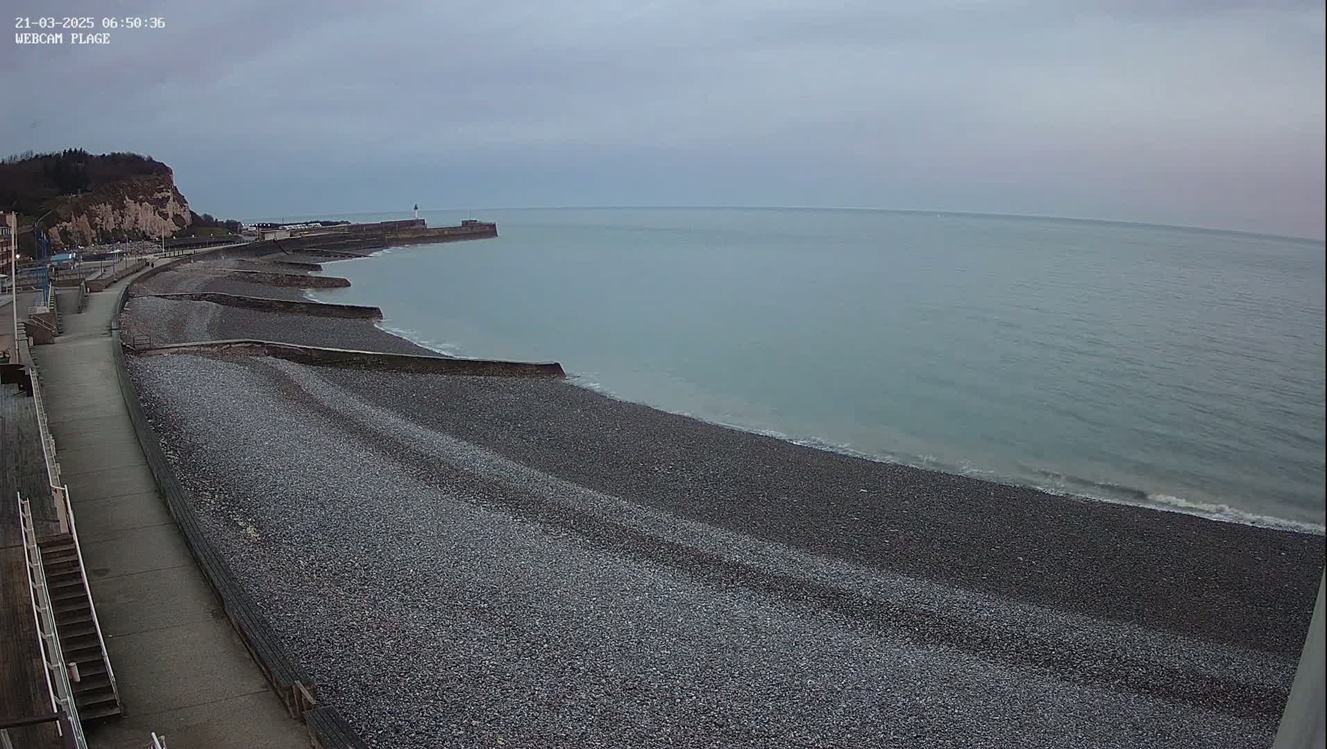 A pebble beach curves alongside a paved walkway, leading to a rocky cliff face under a cloudy sky, with calm, light-grey water visible in the distance.