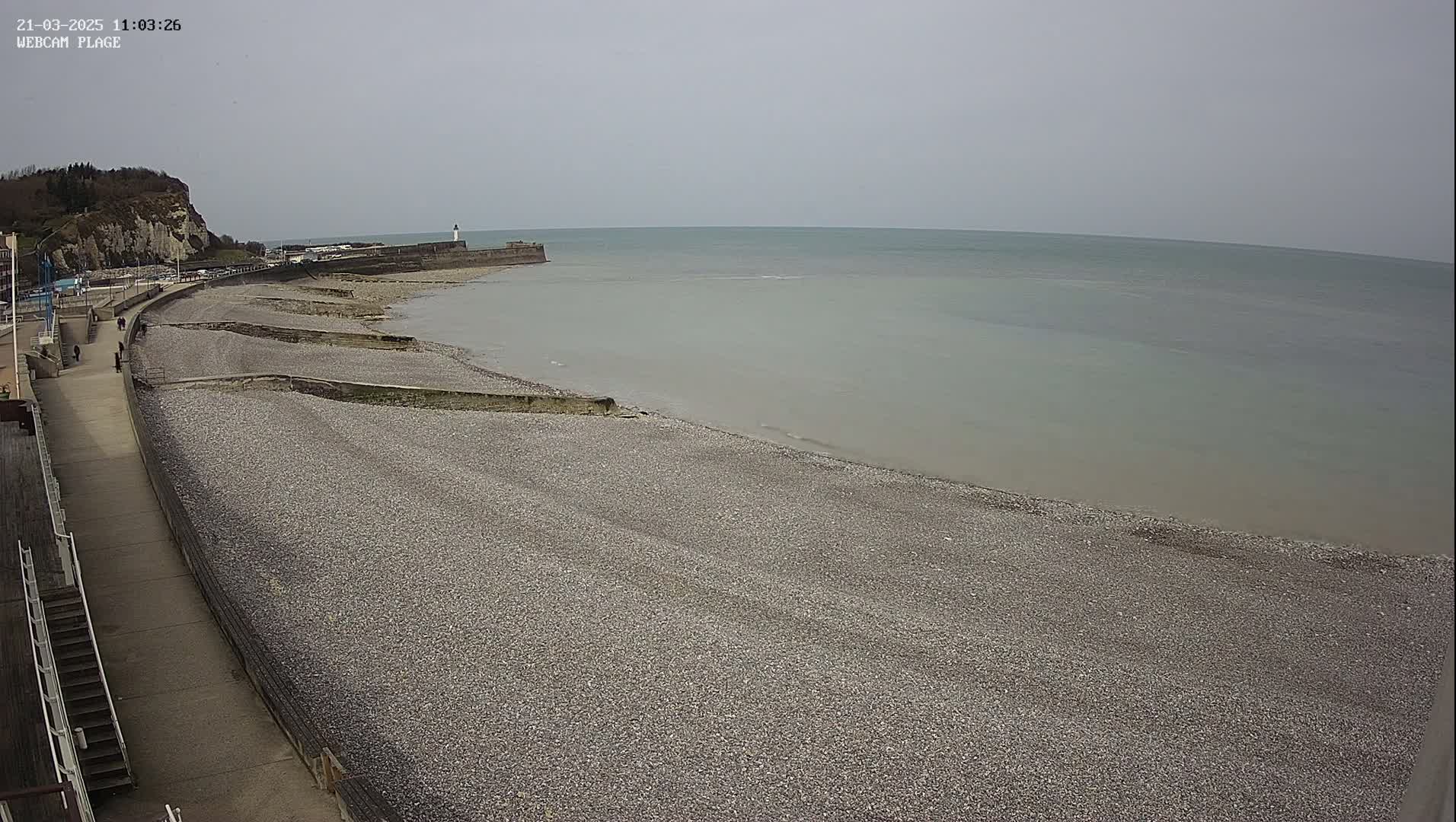 A pebble beach stretches out to a calm, gray sea under an overcast sky, with a walkway and a distant lighthouse visible.