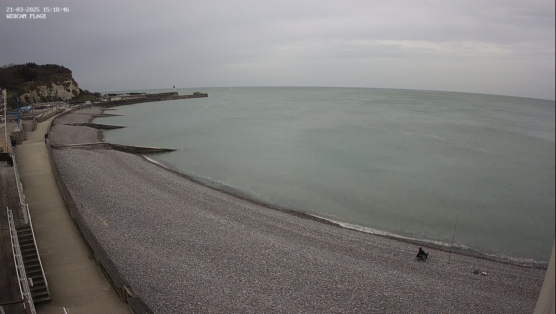 A person sits fishing on a pebble beach next to a calm, gray sea under an overcast sky.