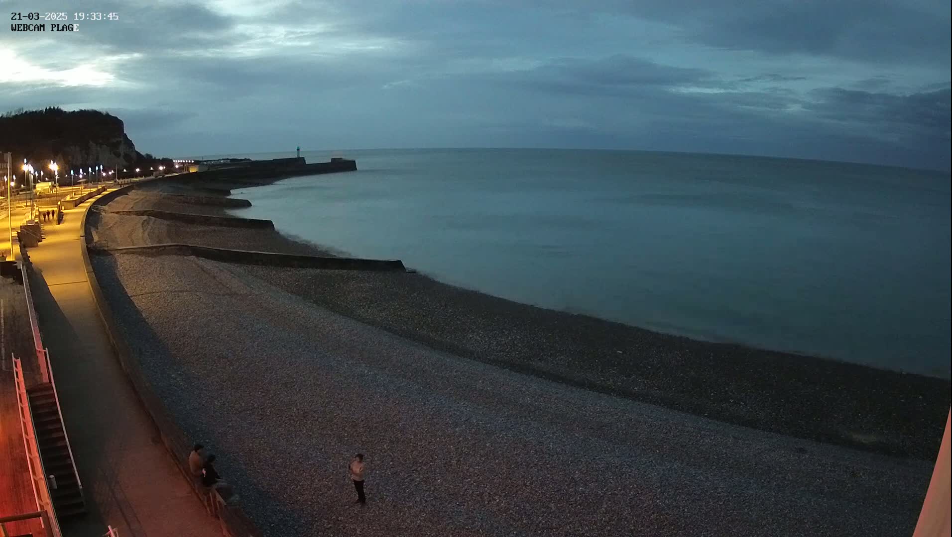 A pebble beach and a paved walkway are illuminated at dusk under a cloudy sky, with a calm sea stretching to the horizon.
