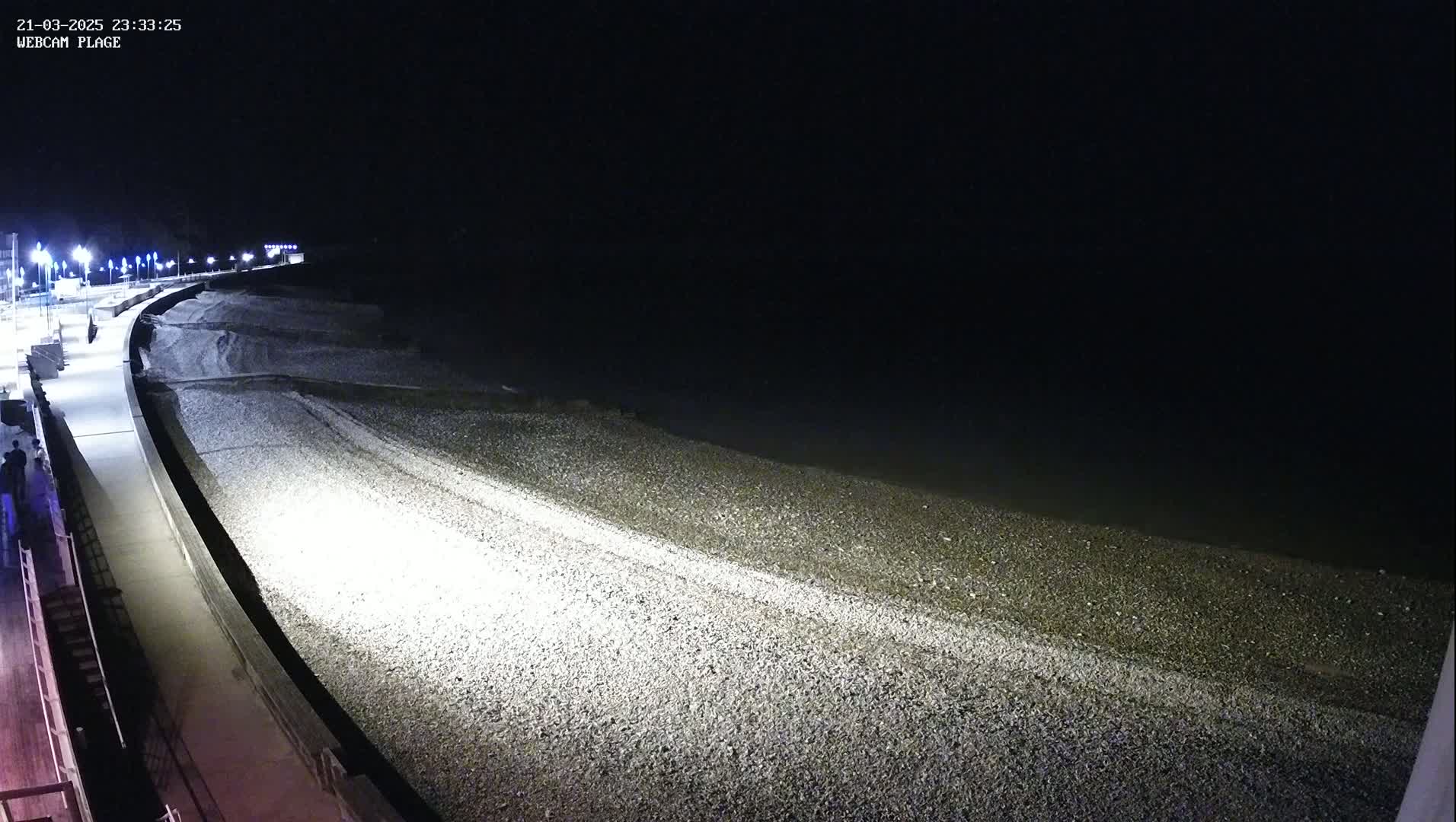 A nighttime view of a pebble beach and a paved walkway, illuminated by artificial lights under a dark sky.