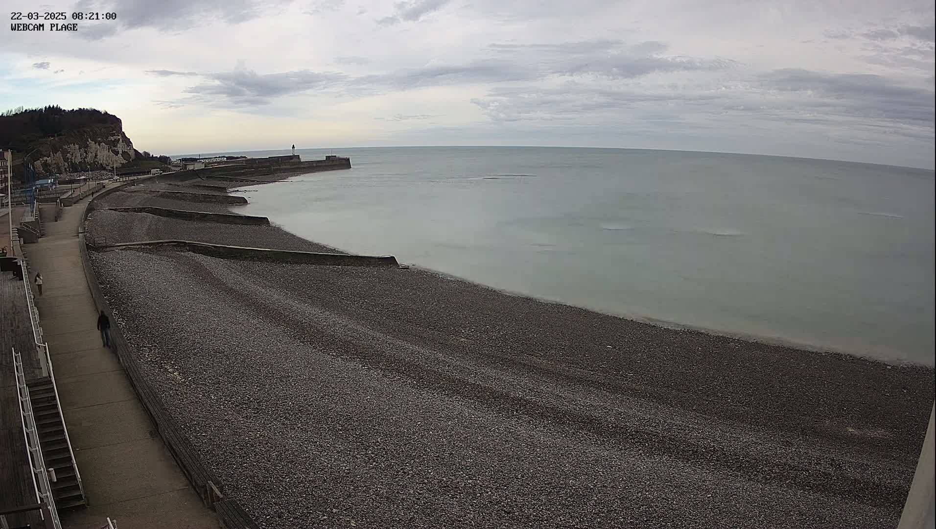 A pebble beach stretches along a calm, gray sea under an overcast sky, next to a paved walkway and a cliff in the distance.