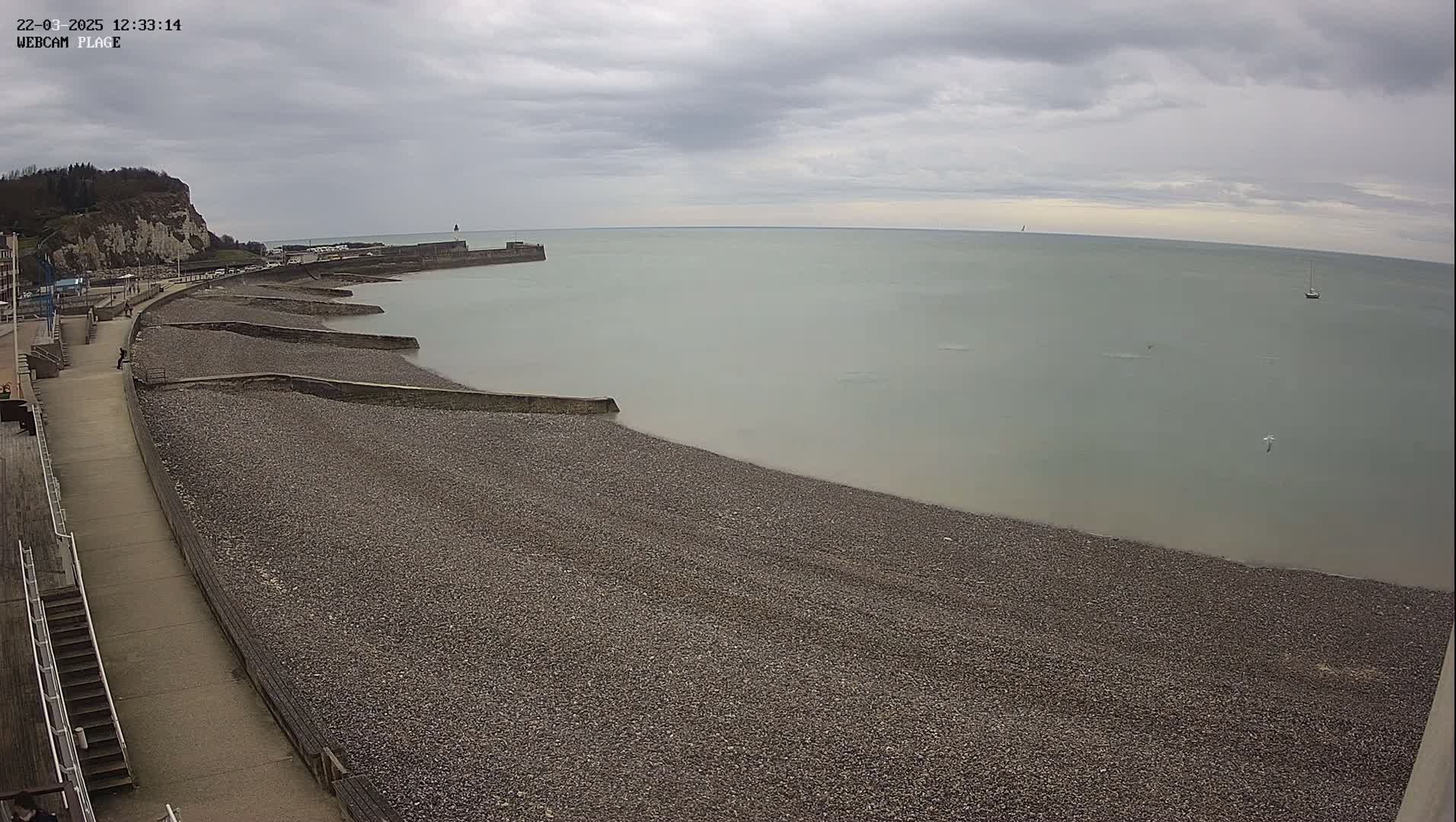 A pebble beach stretches along a calm, gray sea under an overcast sky, with a walkway and low cliffs visible in the background.