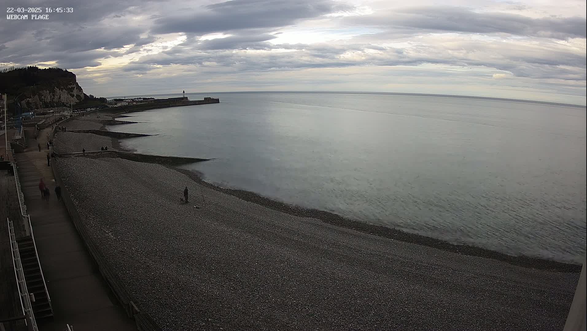 A pebble beach stretches alongside a calm sea under a cloudy sky, with a few people walking along the shore and a pier visible in the distance.