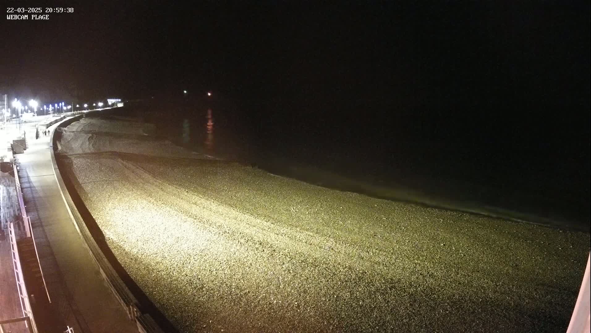 A dark, pebble beach is illuminated at night by artificial lights, with a calm sea gently lapping the shore.