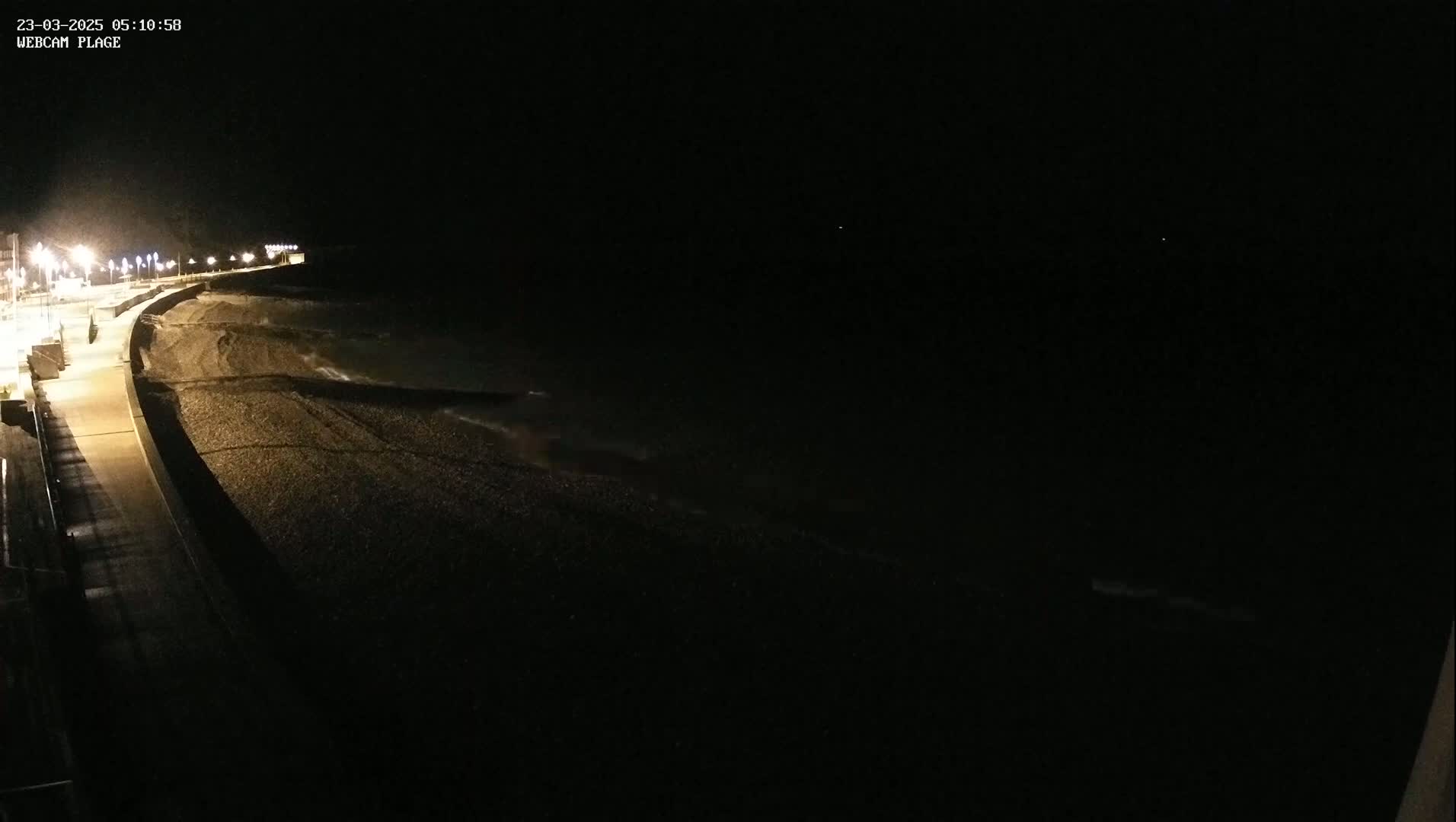 A dark, nighttime view of a pebble beach with gentle waves lapping the shore, illuminated by streetlights along a paved walkway.