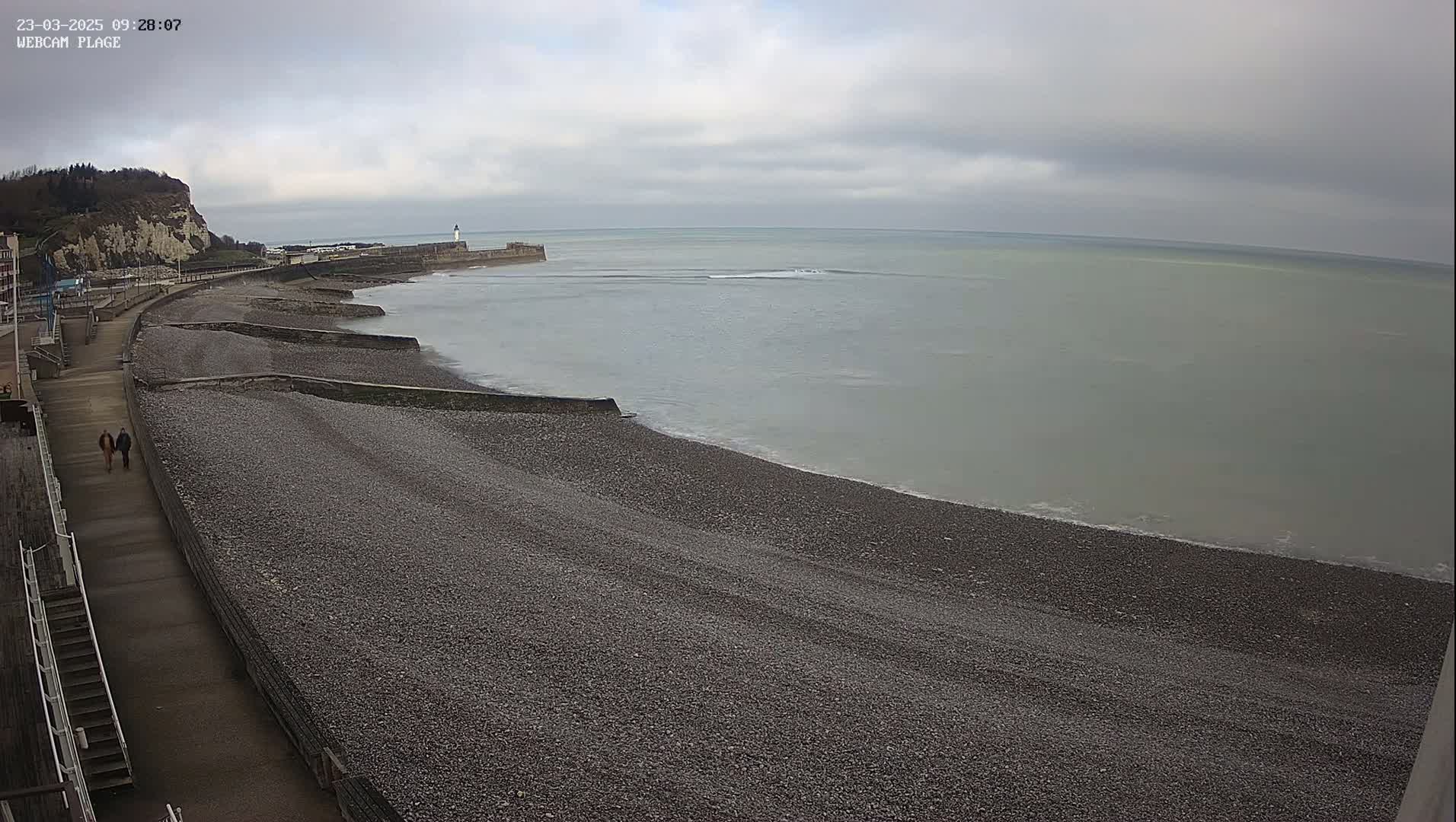 A pebble beach stretches along a calm, gray sea under an overcast sky, with a walkway and two people walking along the shore, near a cliff and a harbor.