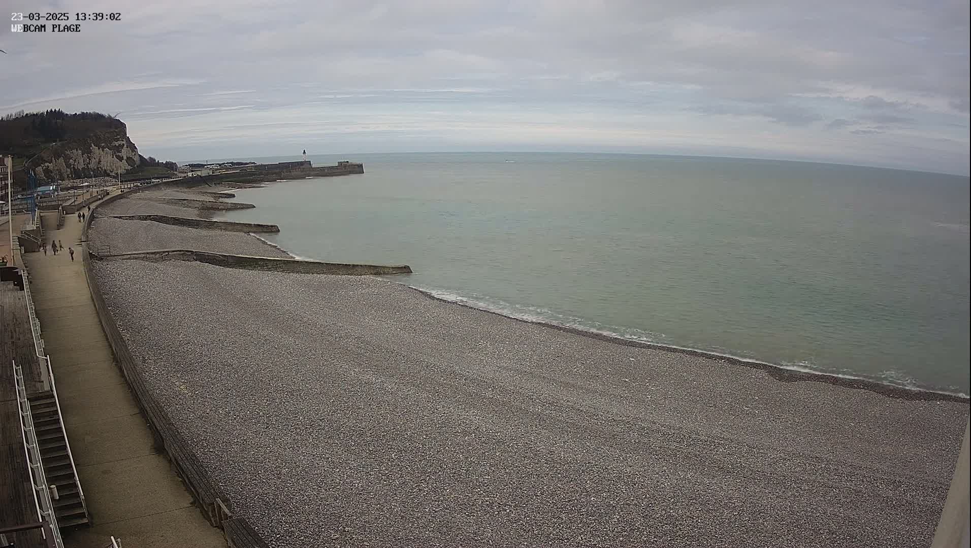 A pebble beach stretches along a calm, gray sea under an overcast sky, with a walkway and a few people visible near the shore.