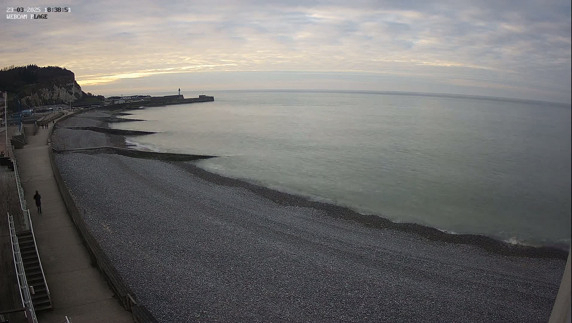 A pebble beach stretches along a calm sea under a cloudy sunset sky, with a walkway and buildings visible in the background.