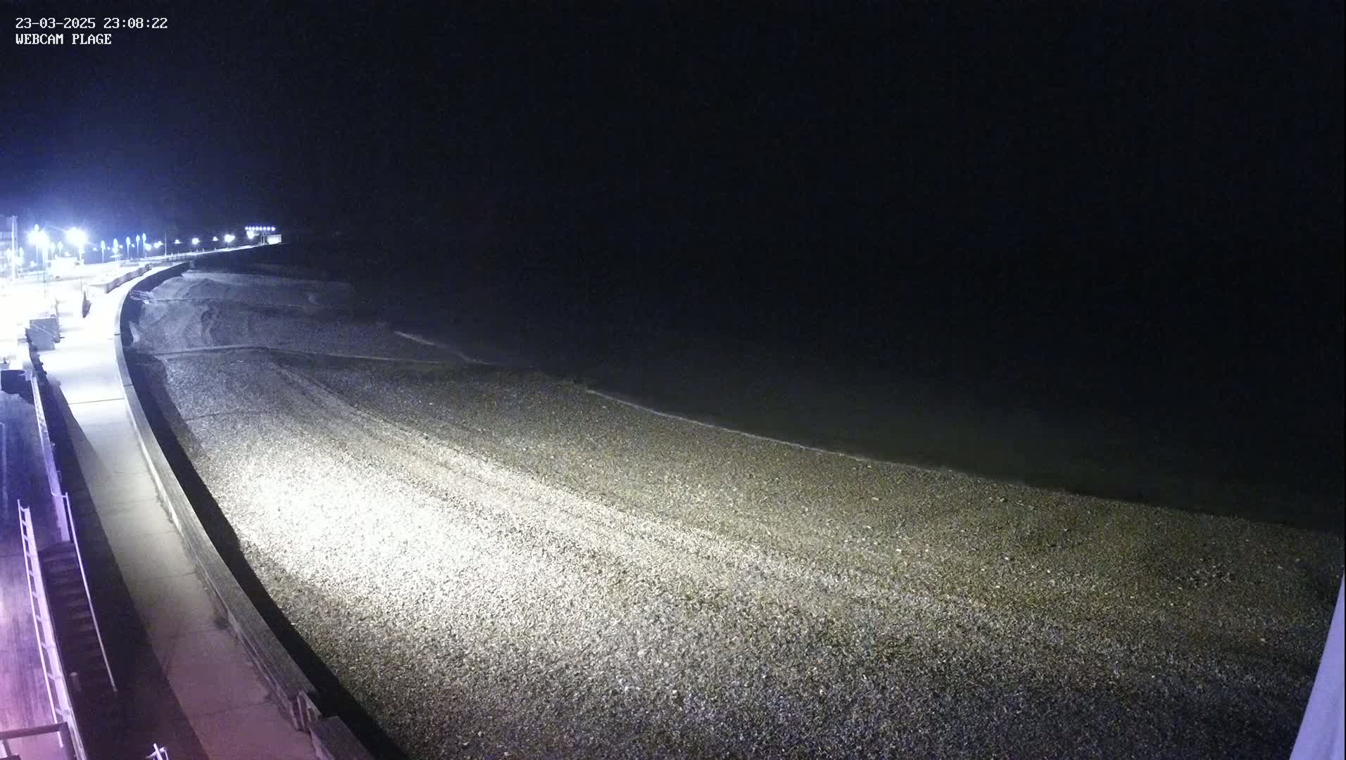 A nighttime view of a pebbled shoreline with gentle waves lapping the shore, illuminated by streetlights along a paved walkway.