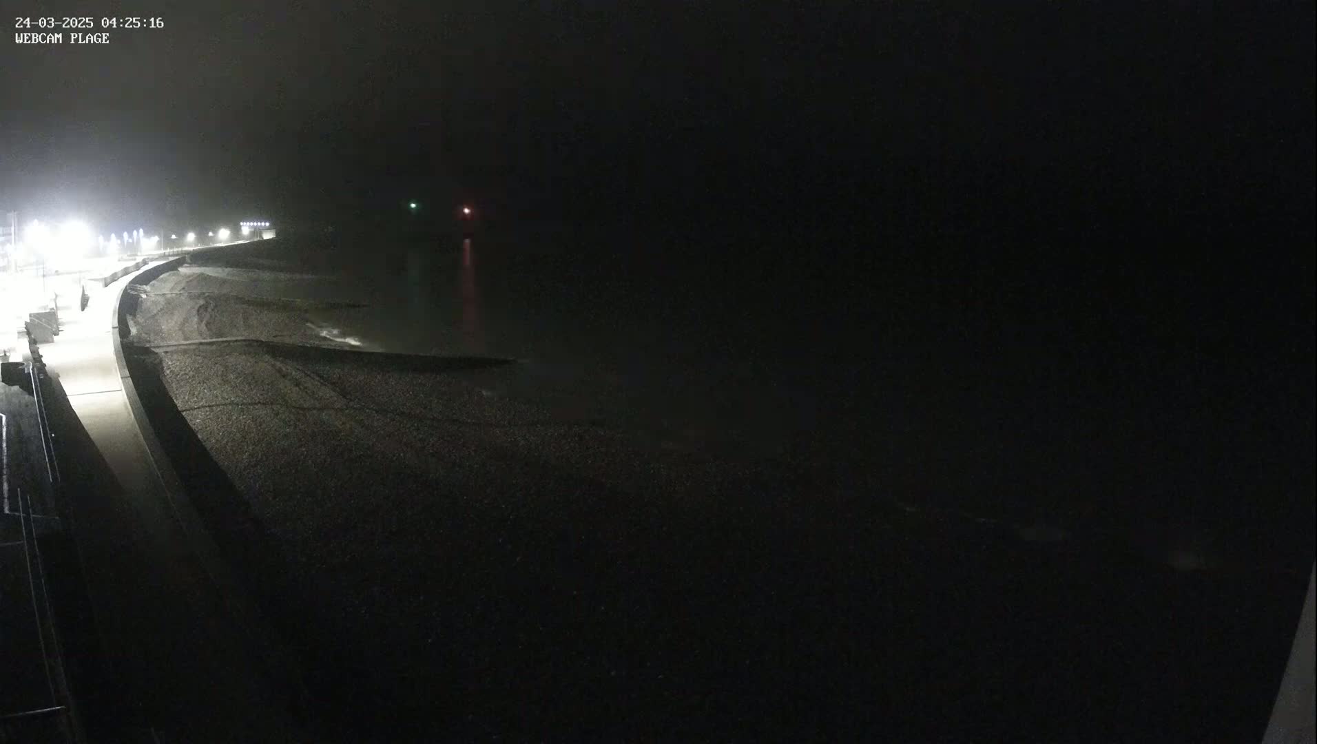 A dark, foggy nighttime scene shows a pebble beach and a paved walkway illuminated by streetlights, with faint lights visible on the water.