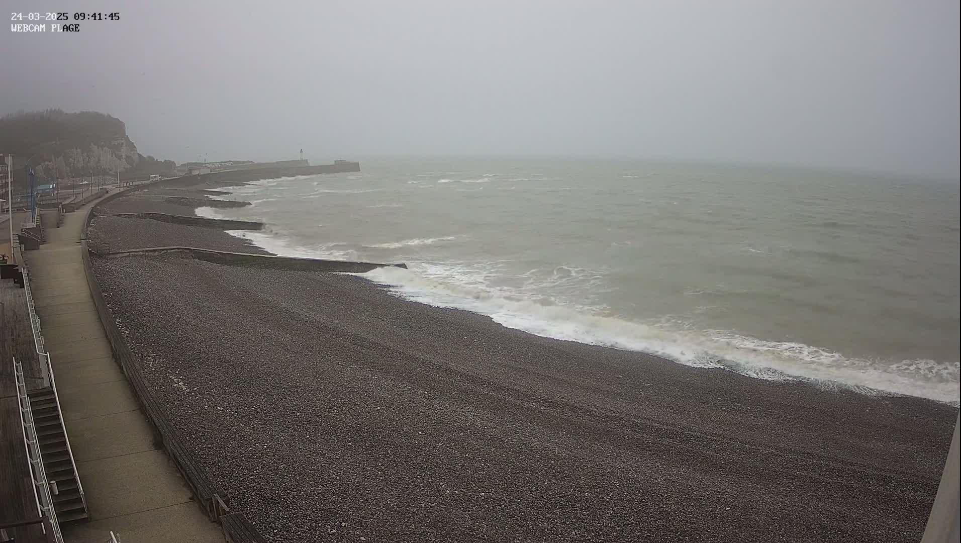 A pebble beach with a walkway and a sea wall is shown, with small waves lapping the shore on an overcast day.