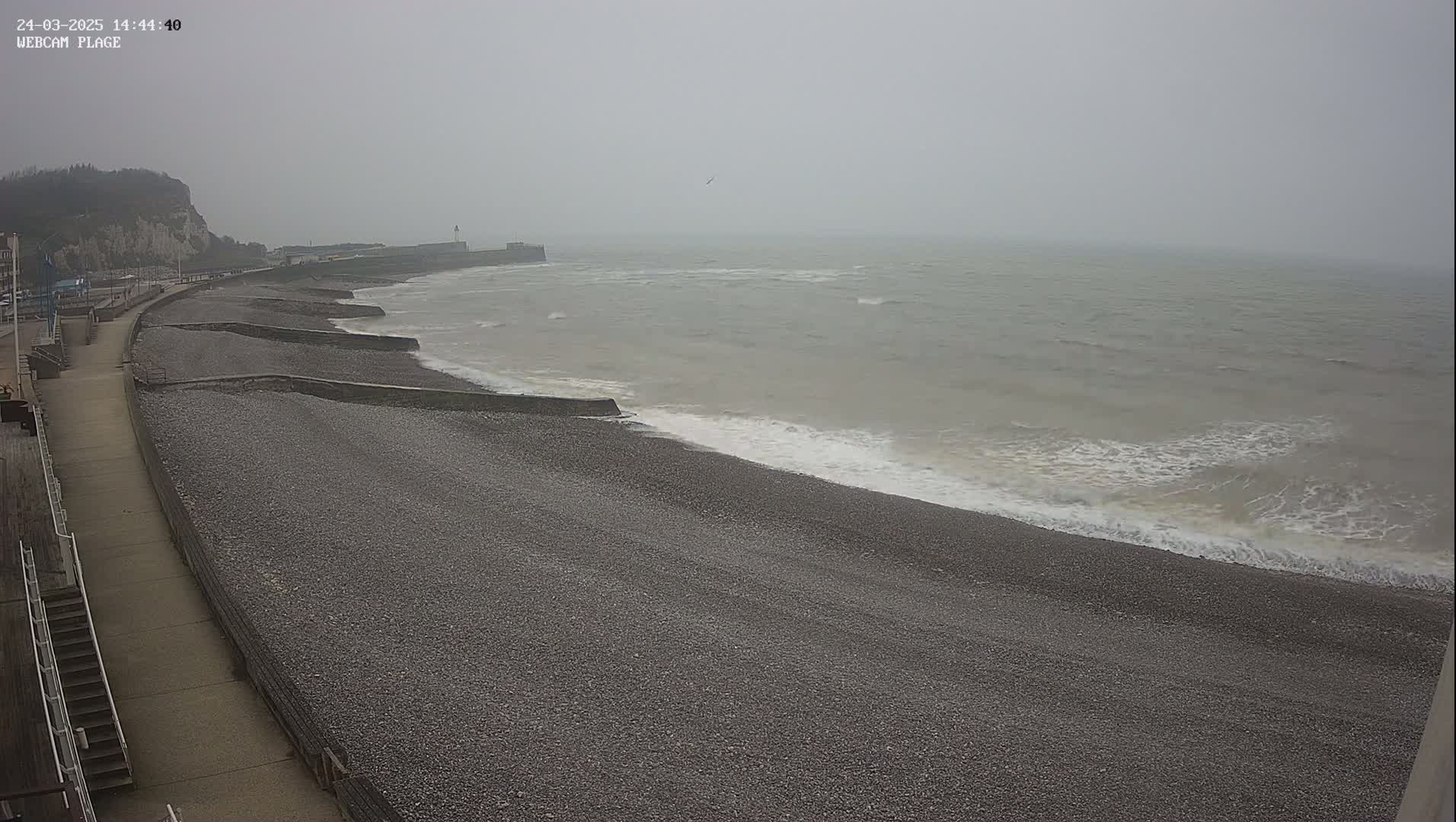 A pebble beach with a walkway and sea wall stretches alongside a grey, choppy sea under an overcast sky.