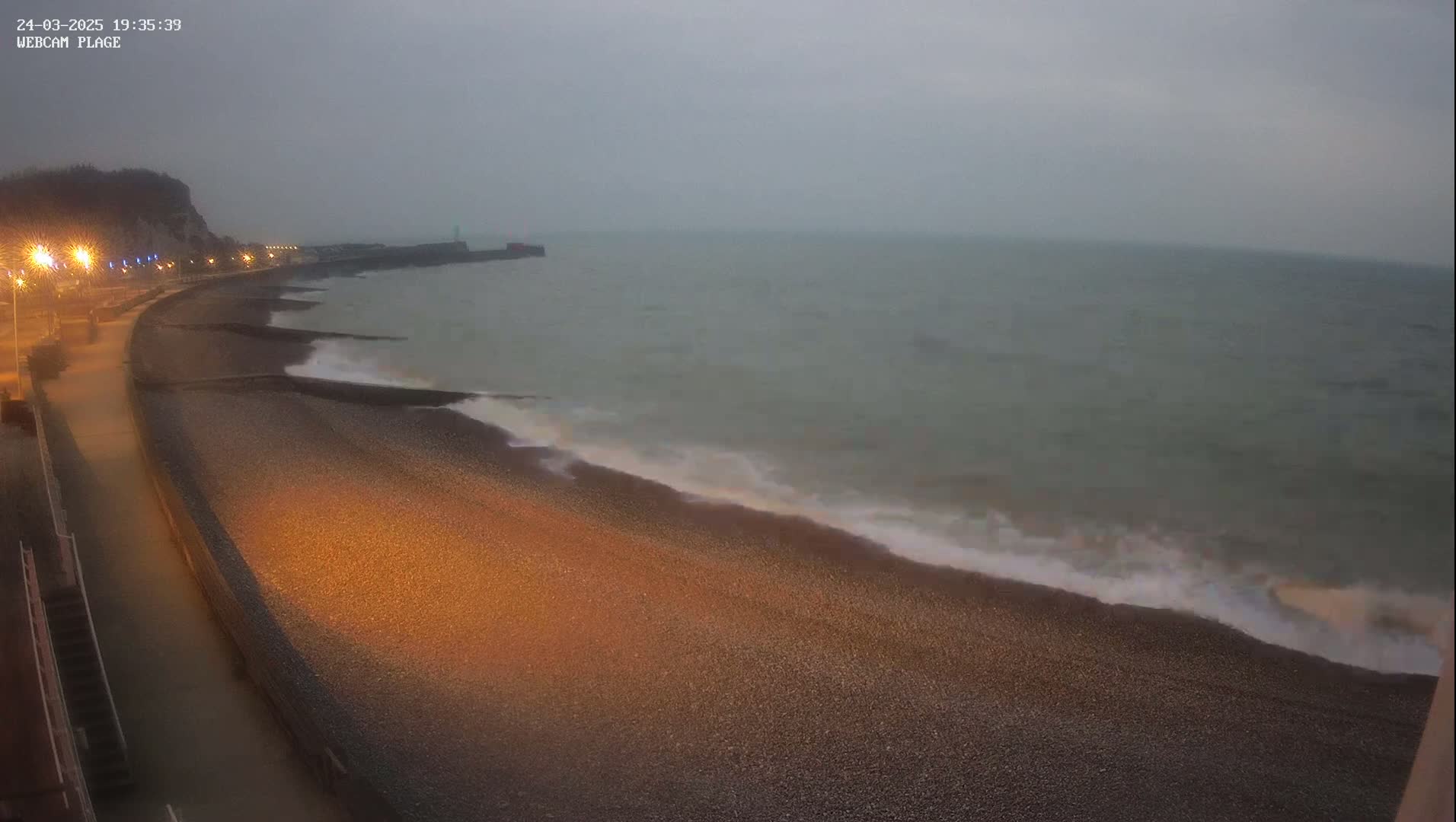 A dark, pebble beach is gently lapped by waves under a cloudy, overcast sky at dusk, with lit pathways visible along a cliffside.